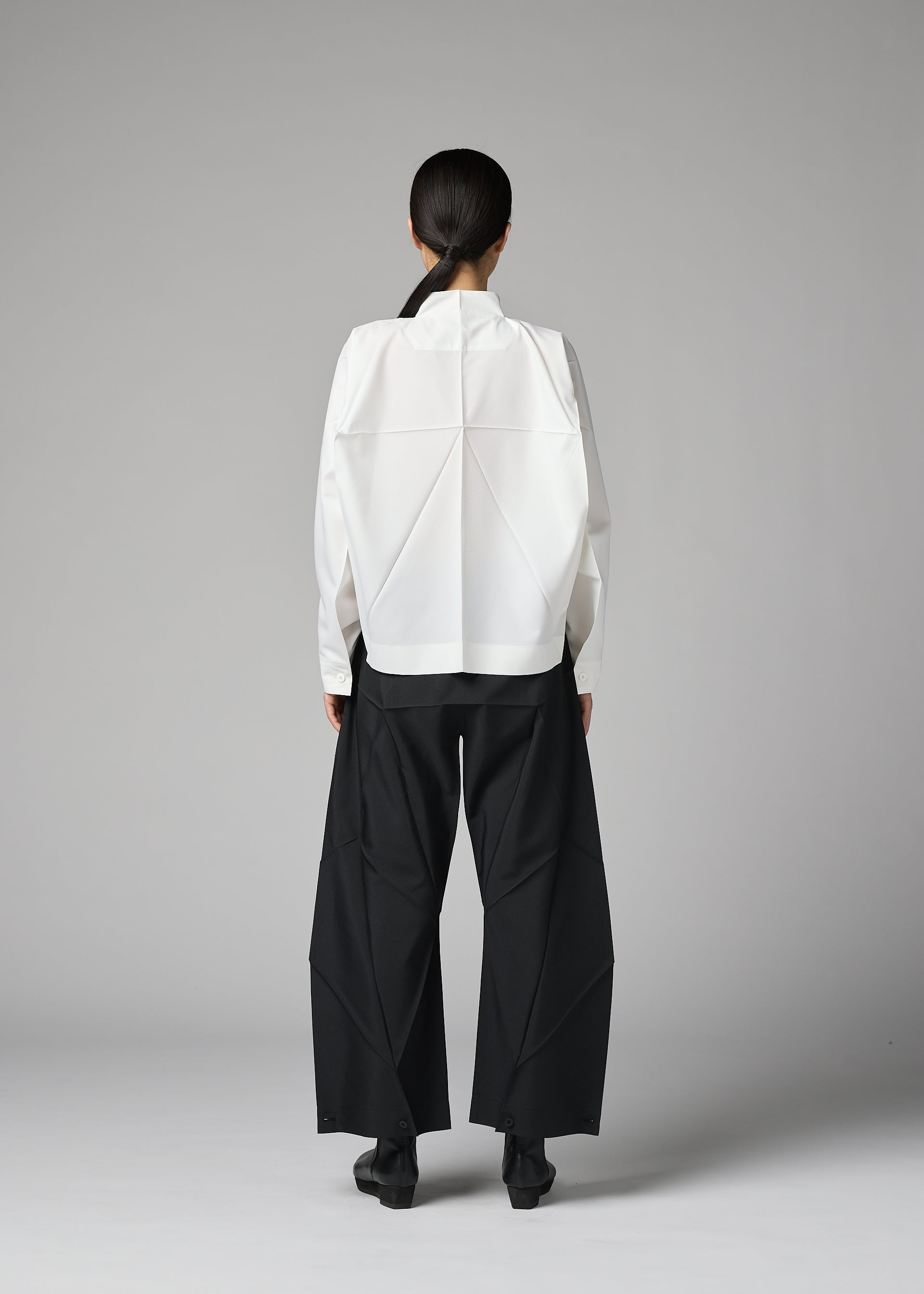 Zoomed view A person stands facing away, wearing the 132 5. ISSEY MIYAKE F-SH-2 shirt featuring geometric folds and wide-leg black pants. Their hair is tied back, set against a plain gray background.