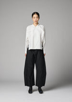 A person stands against a gray background wearing the F-SH-2 shirt by 132 5. ISSEY MIYAKE, featuring an origami-inspired folding technique in white, paired with wide black pleated pants and black shoes. Their hair is pulled back, and they are facing forward with a neutral expression.