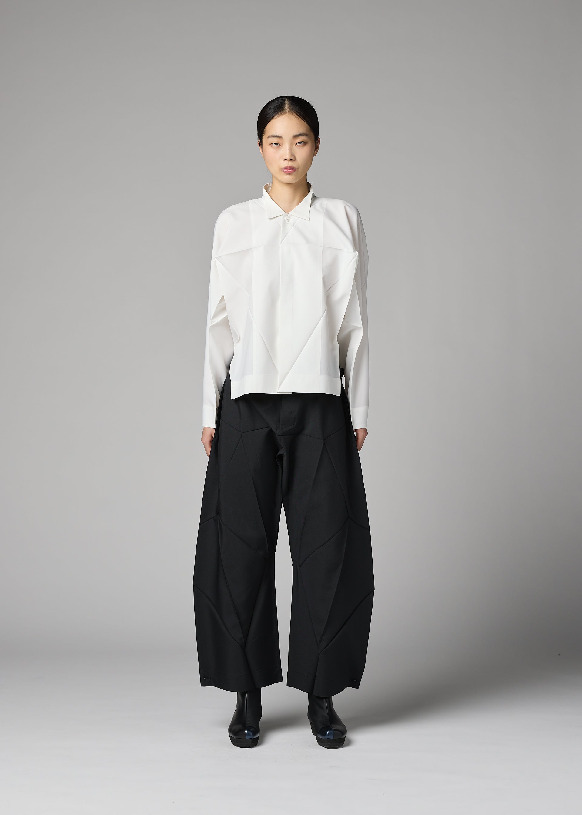 Zoomed view A person stands against a gray background wearing the F-SH-2 shirt by 132 5. ISSEY MIYAKE, featuring an origami-inspired folding technique in white, paired with wide black pleated pants and black shoes. Their hair is pulled back, and they are facing forward with a neutral expression.
