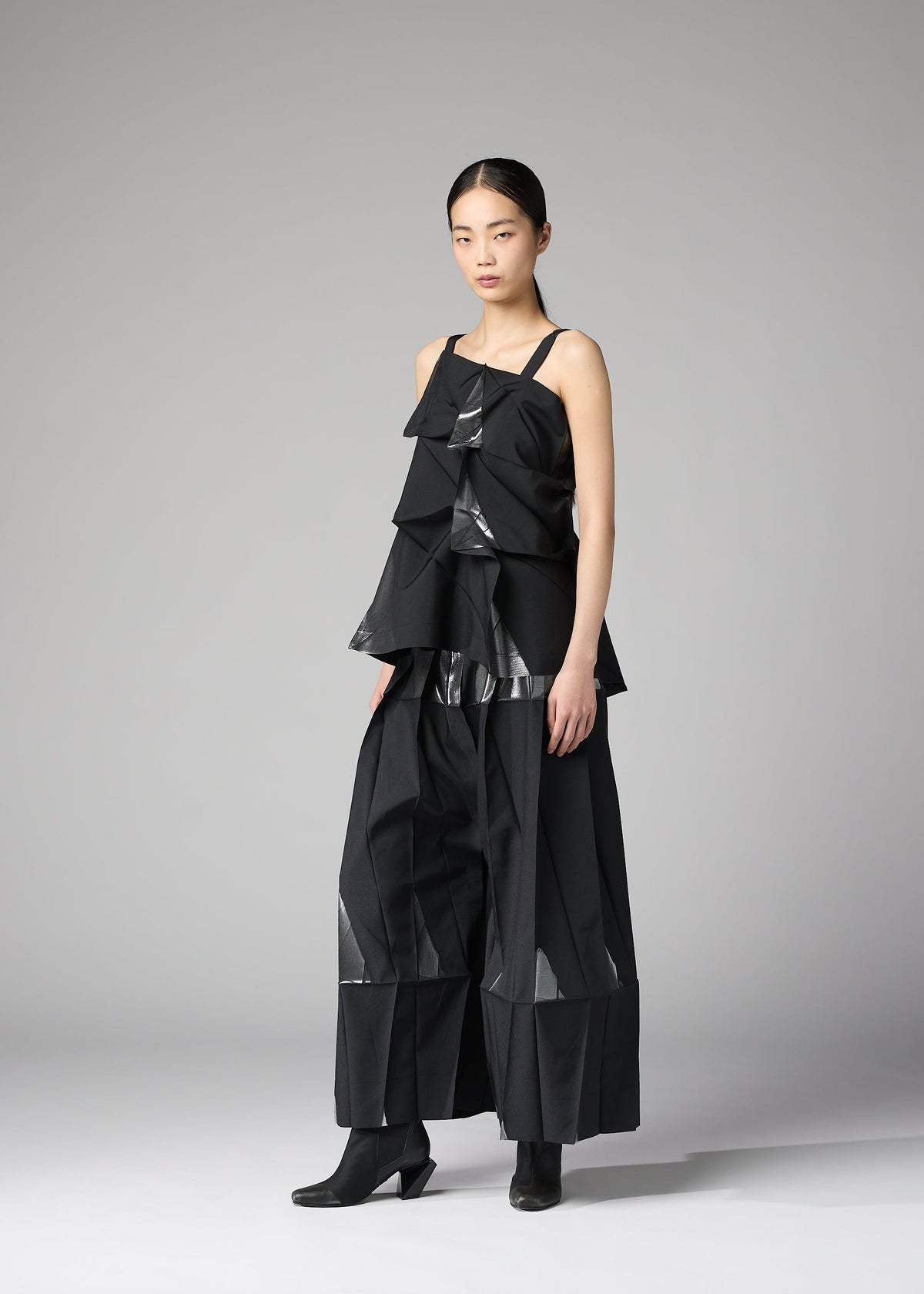 A woman poses against a plain gray background wearing the No.1-1 set from 132 5. ISSEY MIYAKE— a sleeveless recycled polyester top with angular, layered panels forming a pyramid shape, paired with matching wide-legged pants and ankle boots. view 6 of 10