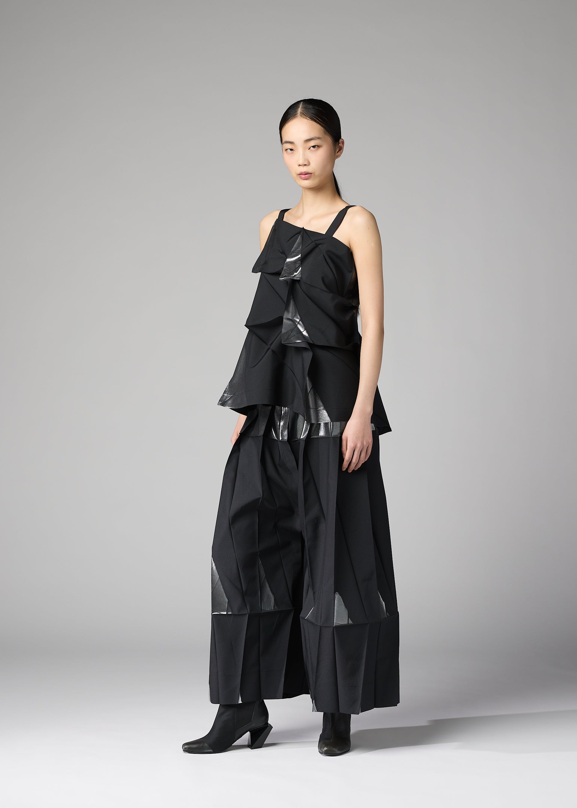 Zoomed view A woman poses against a plain gray background wearing the No.1-1 set from 132 5. ISSEY MIYAKE— a sleeveless recycled polyester top with angular, layered panels forming a pyramid shape, paired with matching wide-legged pants and ankle boots.