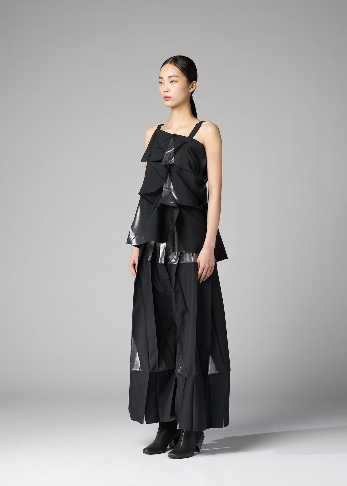 A woman with straight dark hair pulled back stands against a gray background wearing 132 5. ISSEY MIYAKE's No.1-1: a layered black recycled polyester top and wide-leg pants with shiny geometric accents, paired with black ankle boots. view 8 of 10