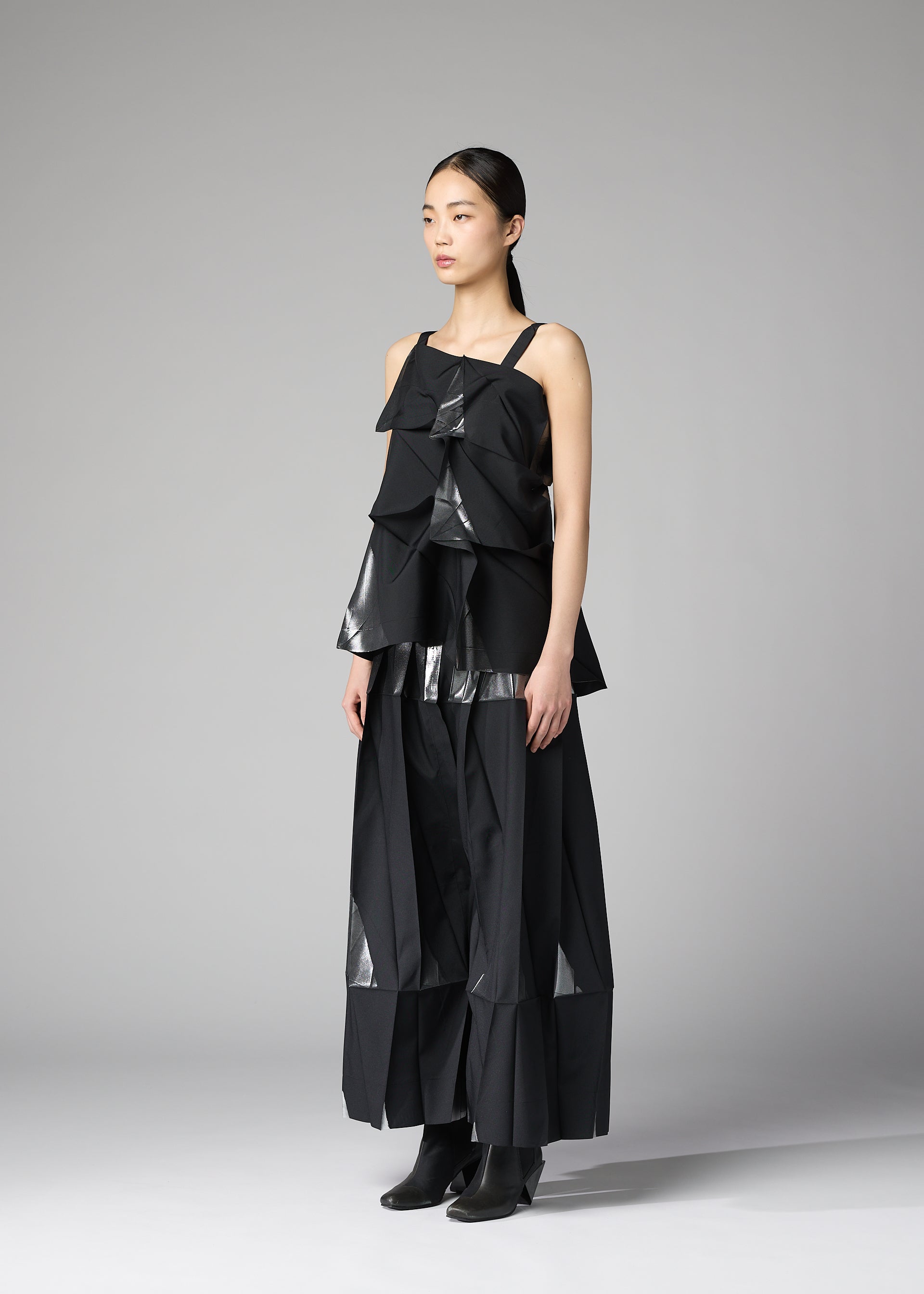 Zoomed view A woman with straight dark hair pulled back stands against a gray background wearing 132 5. ISSEY MIYAKE's No.1-1: a layered black recycled polyester top and wide-leg pants with shiny geometric accents, paired with black ankle boots.
