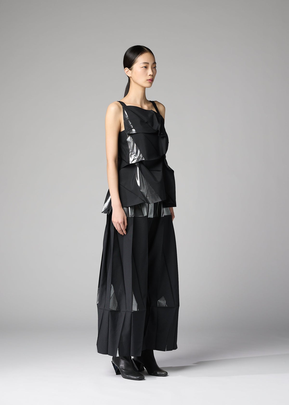 A woman stands in a plain gray studio wearing the No.1-1 set by 132 5. ISSEY MIYAKE—modern black recycled polyester top with layered, angular details and matching wide-leg pants—paired with black ankle boots and sleek, pulled-back hair. view 9 of 10