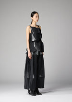 A woman stands in a plain gray studio wearing the No.1-1 set by 132 5. ISSEY MIYAKE—modern black recycled polyester top with layered, angular details and matching wide-leg pants—paired with black ankle boots and sleek, pulled-back hair.