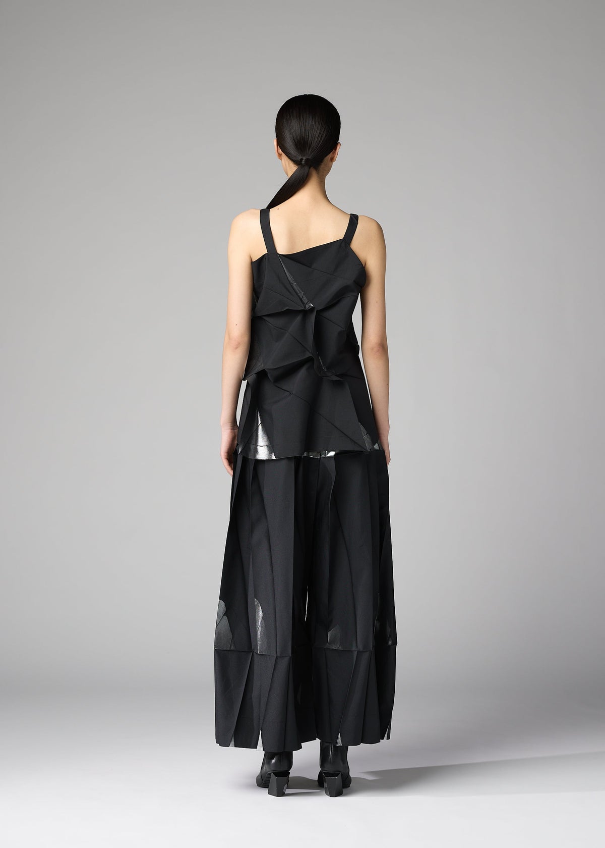 A person stands facing away, wearing the No.1-1 set by 132 5. ISSEY MIYAKE: a geometric black recycled polyester top with asymmetrical folds and wide-leg pants, featuring sheer panels and sculptural shapes against a plain gray background. view 10 of 10