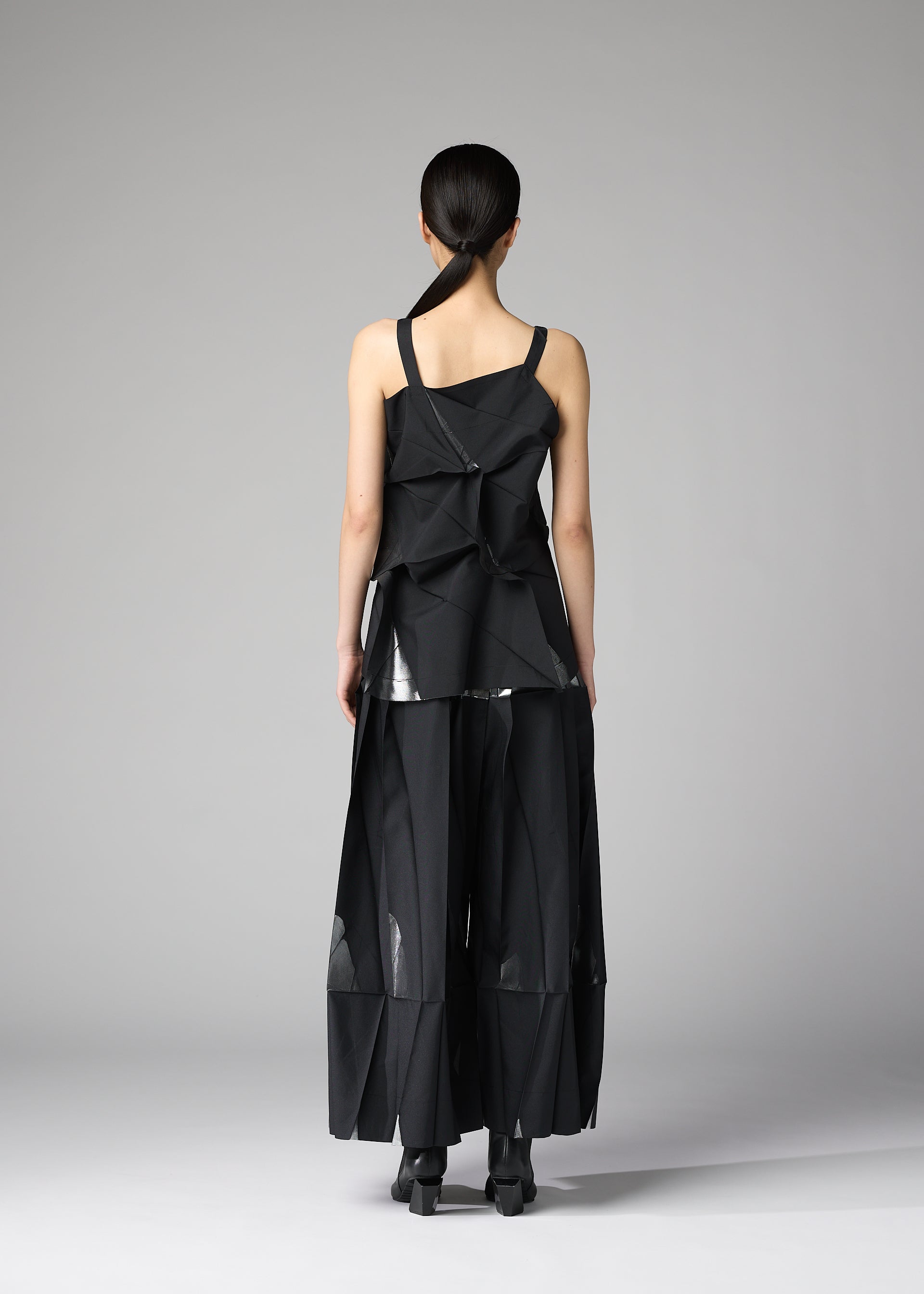 Zoomed view A person stands facing away, wearing the No.1-1 set by 132 5. ISSEY MIYAKE: a geometric black recycled polyester top with asymmetrical folds and wide-leg pants, featuring sheer panels and sculptural shapes against a plain gray background.
