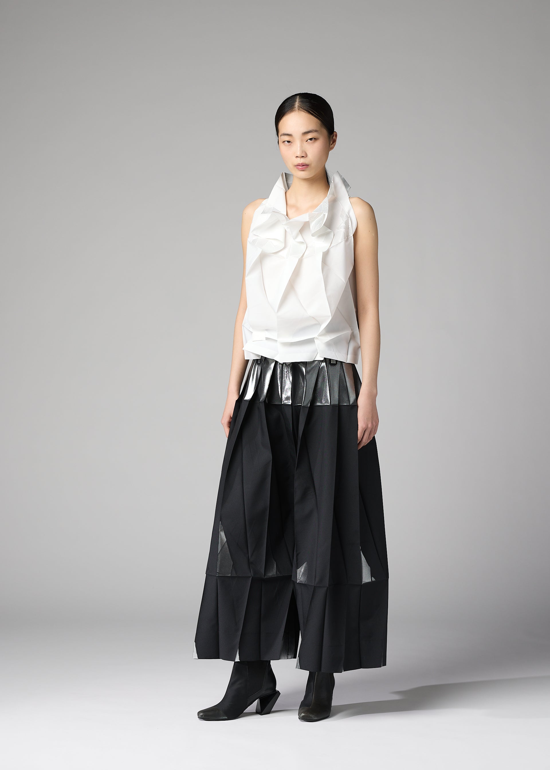 Zoomed view A woman poses against a gray background wearing the No.10-1 top by 132 5. ISSEY MIYAKE, featuring recycled polyester, a sculptural collar, metallic silver pleated accent, wide black culottes with geometric panels, and black ankle boots.