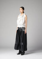 A woman stands in a studio against a plain gray background wearing 132 5. ISSEY MIYAKE's No.10-1: a white sculptural recycled polyester top with large folds, a long black skirt with sheer panels, and black ankle boots. Her straight dark hair is tied back.