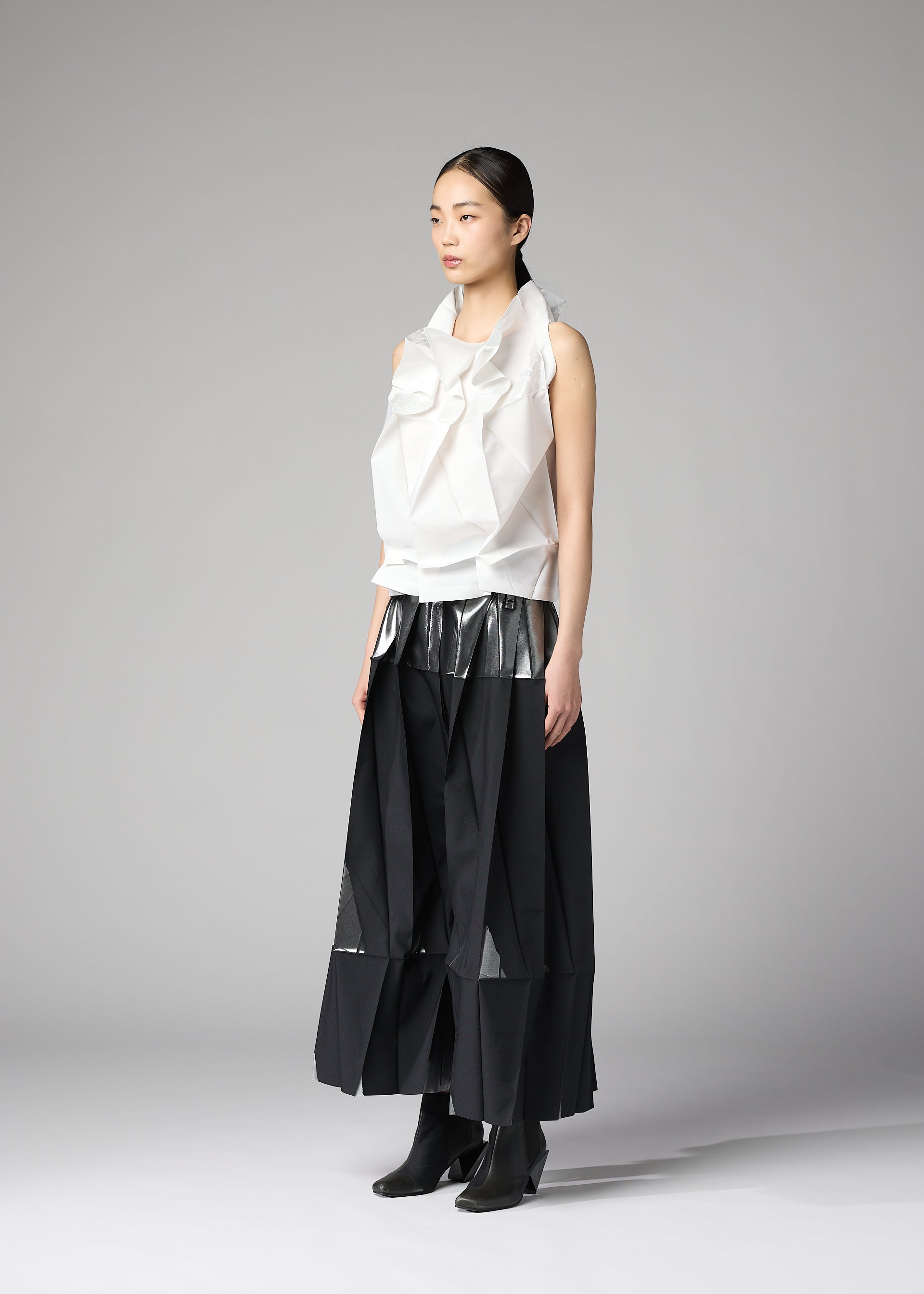 Zoomed view A woman stands in a studio against a plain gray background wearing 132 5. ISSEY MIYAKE's No.10-1: a white sculptural recycled polyester top with large folds, a long black skirt with sheer panels, and black ankle boots. Her straight dark hair is tied back.