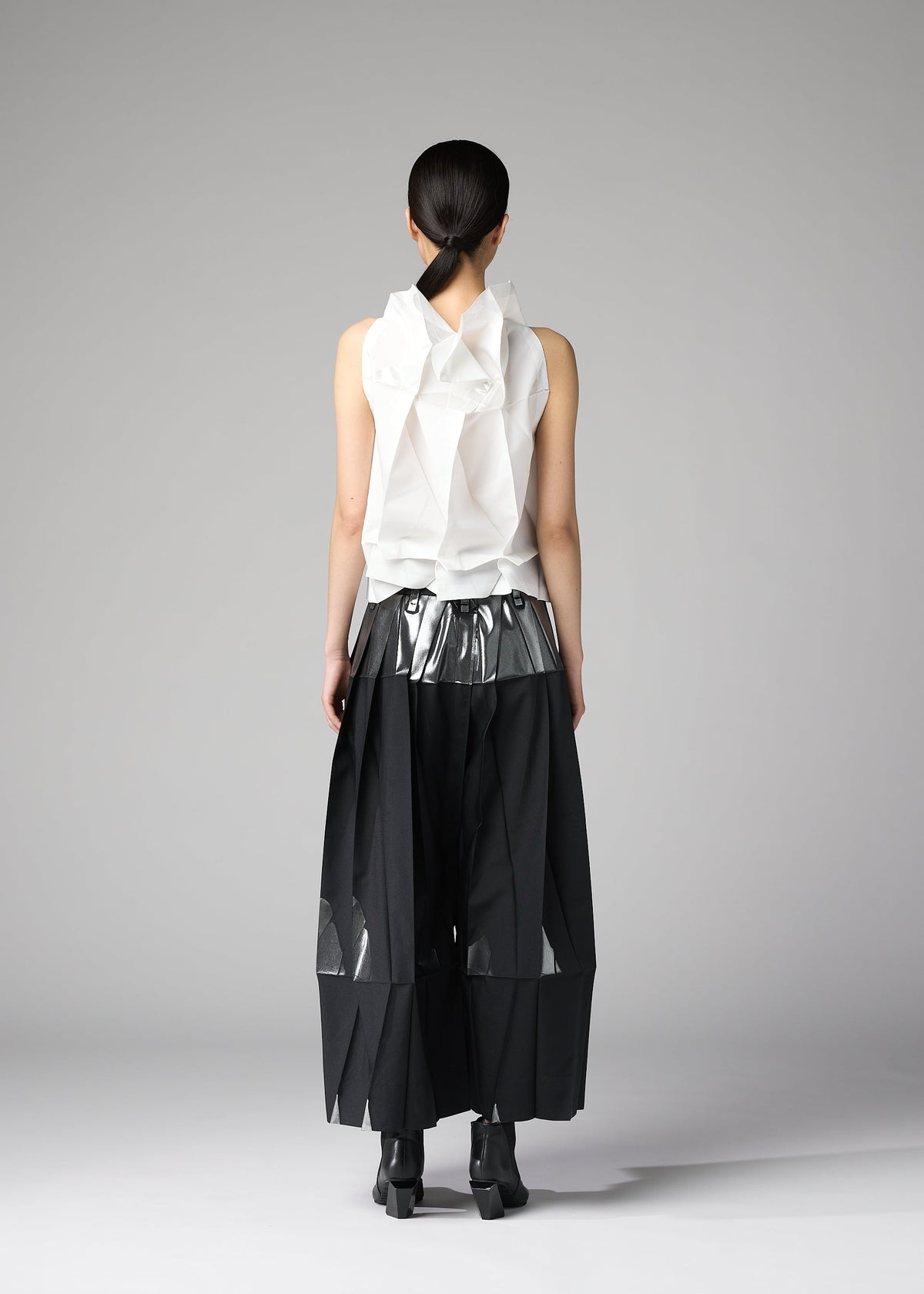A person faces away, wearing 132 5. ISSEY MIYAKE's No.10-1: a sleeveless white foil-detail ruffled top with metallic silver waistband, wide black pleated mesh-panel pants, black ankle boots, and sleek dark hair tied back. view 8 of 8
