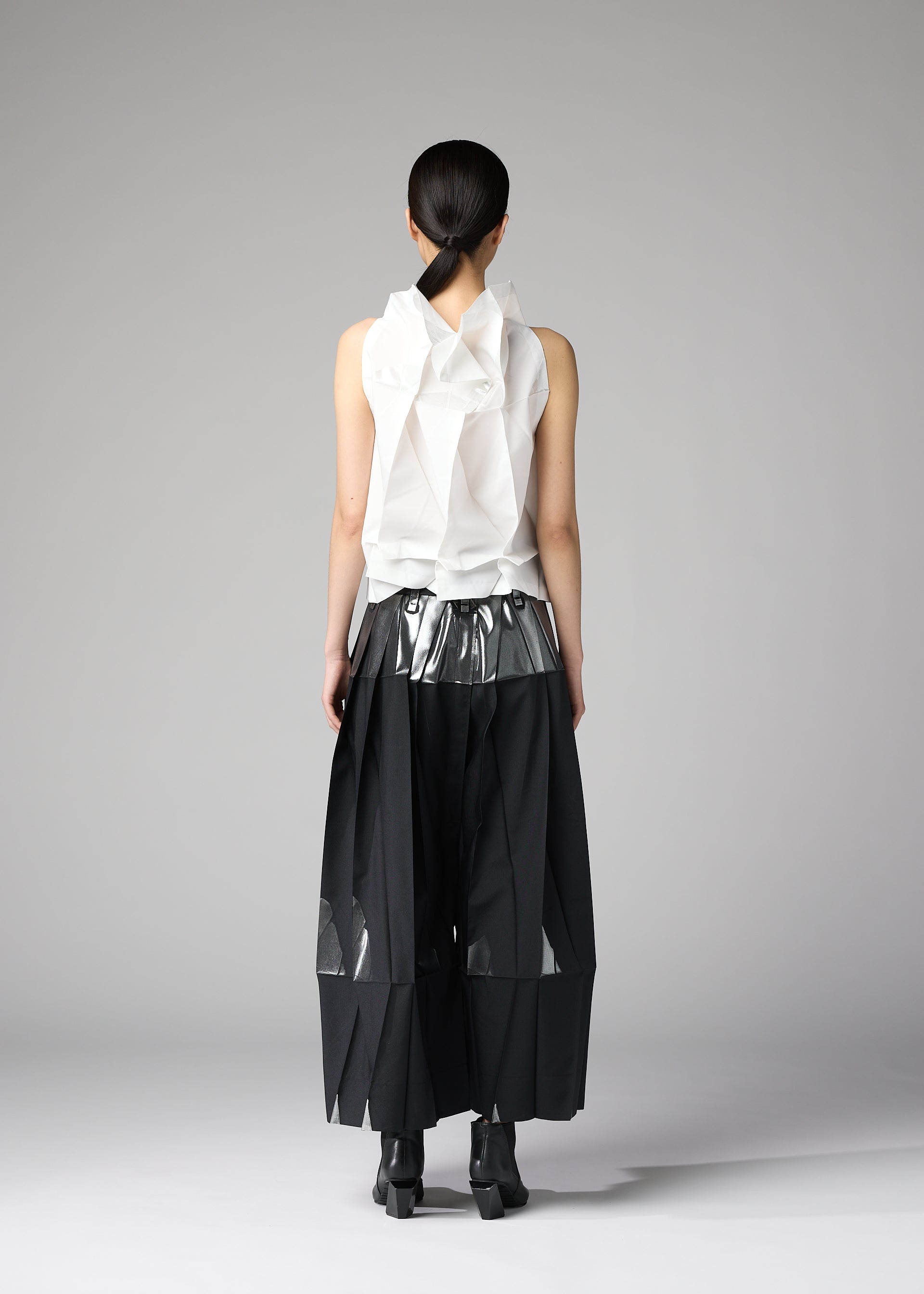 Zoomed view A person faces away, wearing 132 5. ISSEY MIYAKE's No.10-1: a sleeveless white foil-detail ruffled top with metallic silver waistband, wide black pleated mesh-panel pants, black ankle boots, and sleek dark hair tied back.