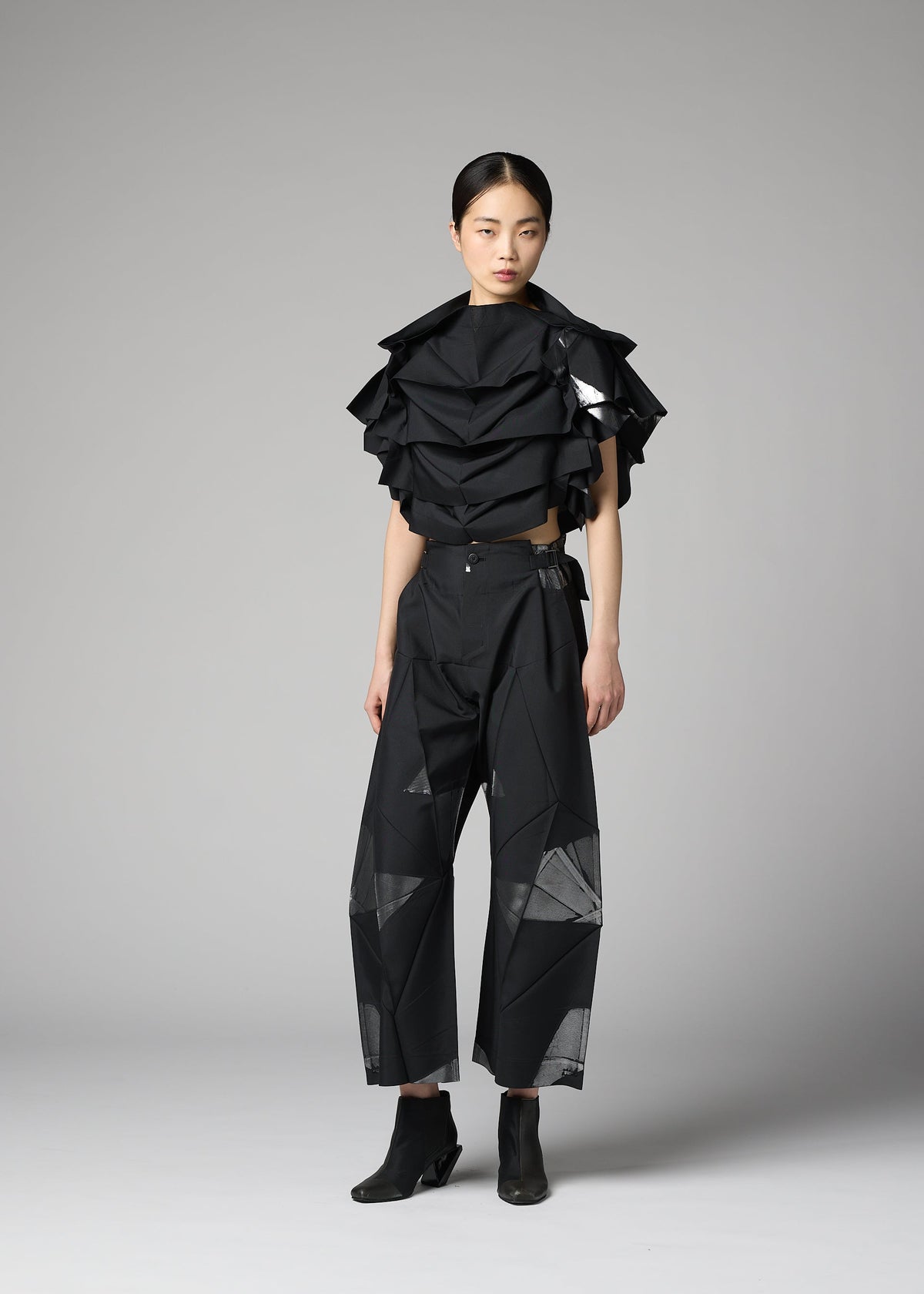 A person stands in a studio wearing the No.10-2 by 132 5. ISSEY MIYAKE—a sculptural black top made from recycled polyester with ruffled layers, paired with black wide-leg pants featuring geometric mesh panels and black ankle boots. The glamorous ensemble is set against a plain gray background. view 6 of 9