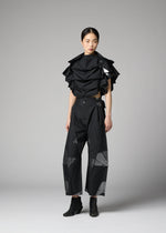 A person stands in a studio wearing the No.10-2 by 132 5. ISSEY MIYAKE—a sculptural black top made from recycled polyester with ruffled layers, paired with black wide-leg pants featuring geometric mesh panels and black ankle boots. The glamorous ensemble is set against a plain gray background.