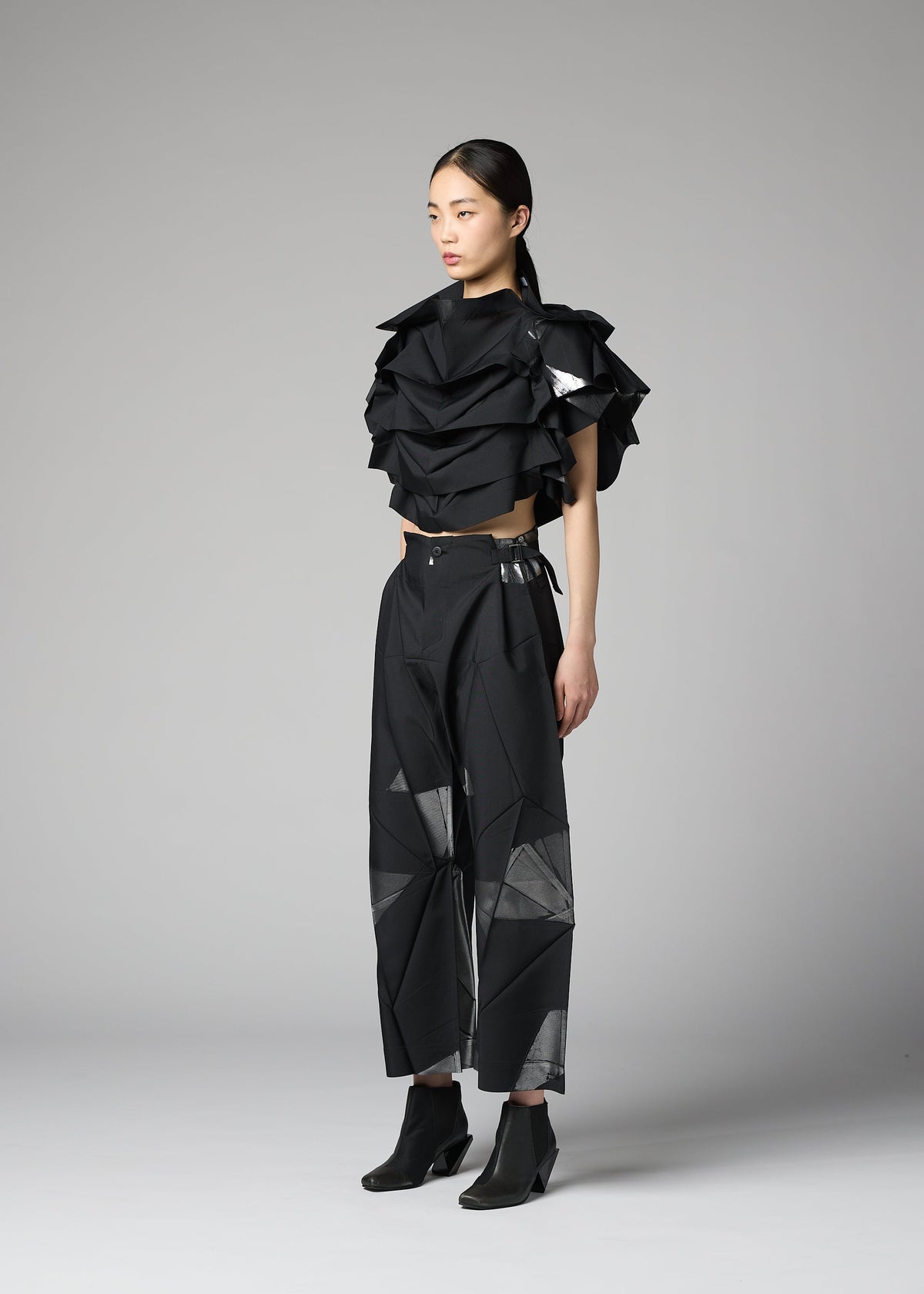 Dressed in the No.10-2 by 132 5. ISSEY MIYAKE, a person stands against a gray background wearing a glamorous black geometric ruffled top made from recycled polyester and wide-leg pants with angular patterns, completed with black ankle boots. view 7 of 9