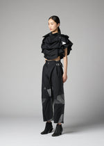 Dressed in the No.10-2 by 132 5. ISSEY MIYAKE, a person stands against a gray background wearing a glamorous black geometric ruffled top made from recycled polyester and wide-leg pants with angular patterns, completed with black ankle boots.