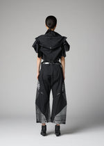 A person stands facing away, dressed in the 132 5. ISSEY MIYAKE No.10-2: a black sculptural shirt with angular layers crafted from recycled polyester, complemented by wide black pants with geometric mesh details and paired with black heeled shoes, against a plain gray background.