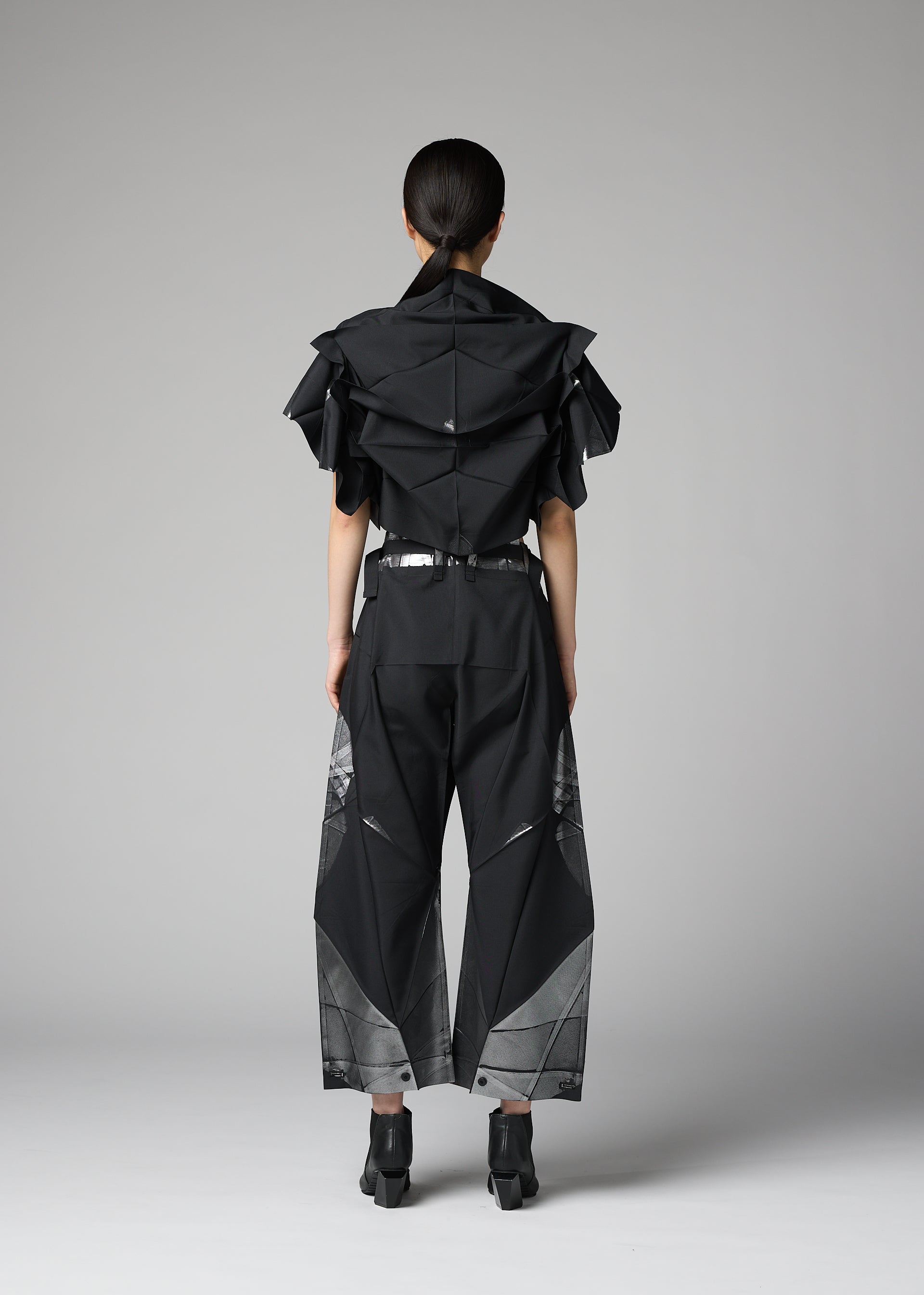 Zoomed view A person stands facing away, dressed in the 132 5. ISSEY MIYAKE No.10-2: a black sculptural shirt with angular layers crafted from recycled polyester, complemented by wide black pants with geometric mesh details and paired with black heeled shoes, against a plain gray background.