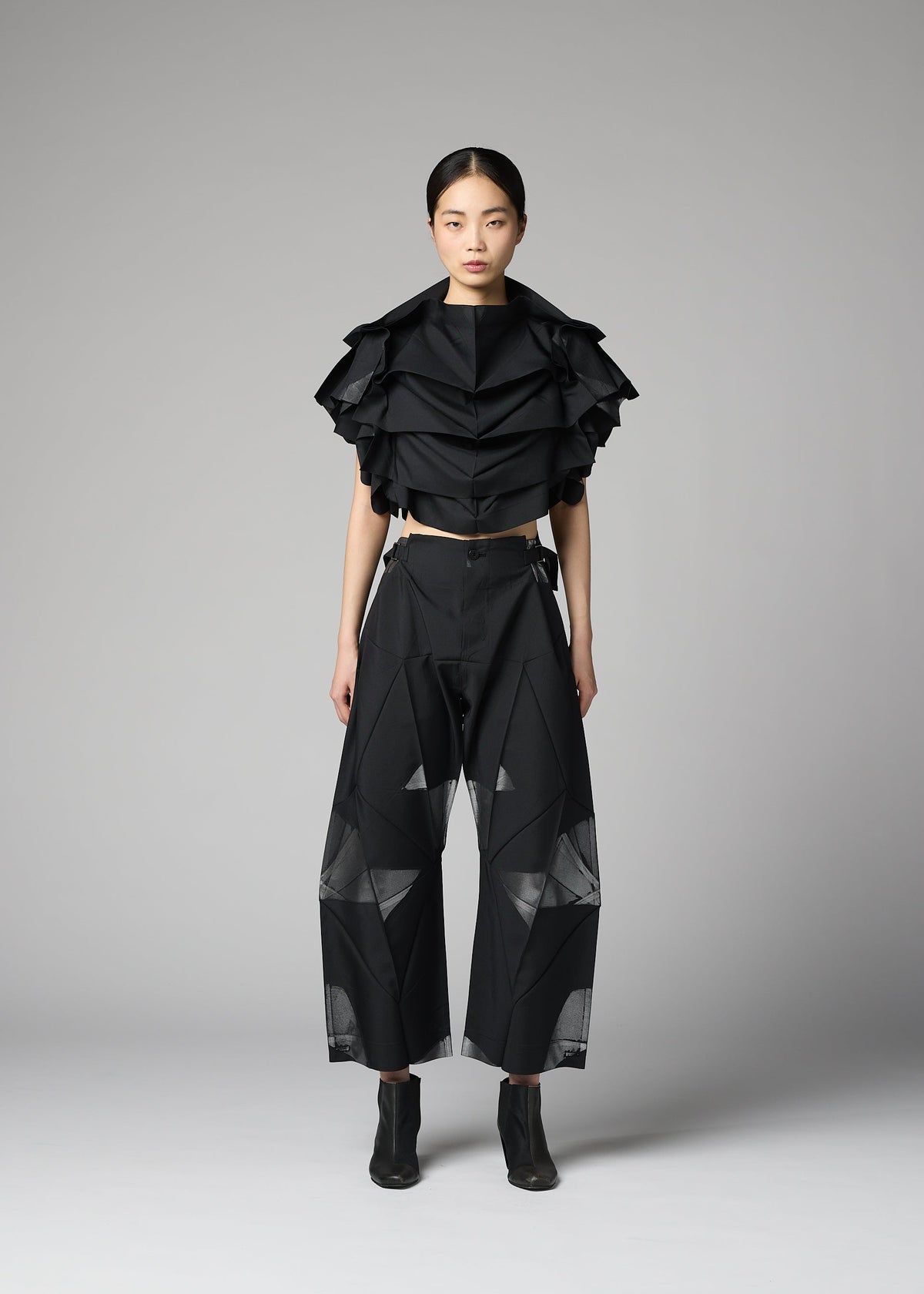 A woman stands against a plain background wearing the 132 5. ISSEY MIYAKE No.10-2 ensemble: a glamorous black ruffled crop top and wide-leg black pants with sheer geometric panels, crafted from recycled polyester, paired with black ankle boots. view 9 of 9