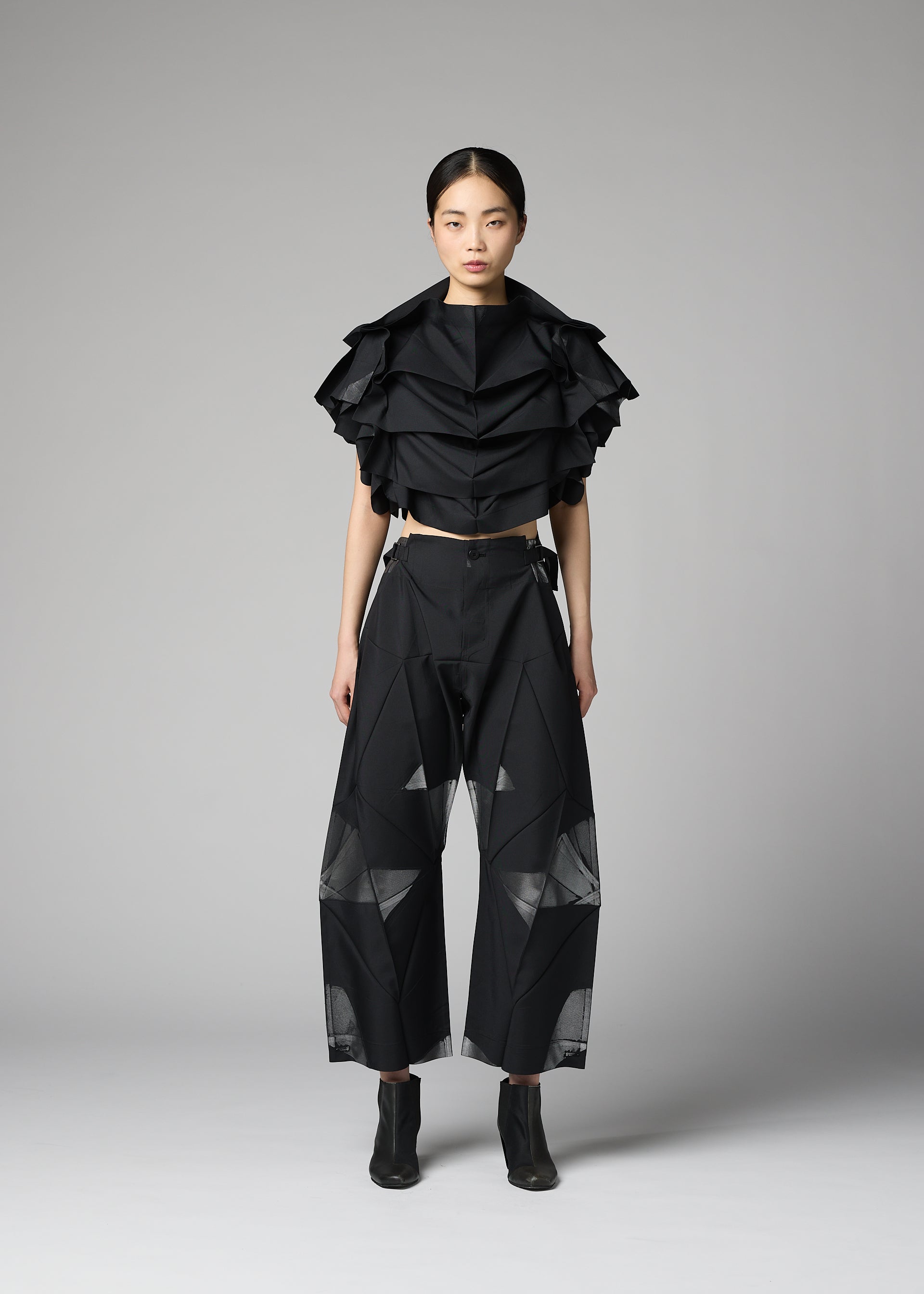 Zoomed view A woman stands against a plain background wearing the 132 5. ISSEY MIYAKE No.10-2 ensemble: a glamorous black ruffled crop top and wide-leg black pants with sheer geometric panels, crafted from recycled polyester, paired with black ankle boots.