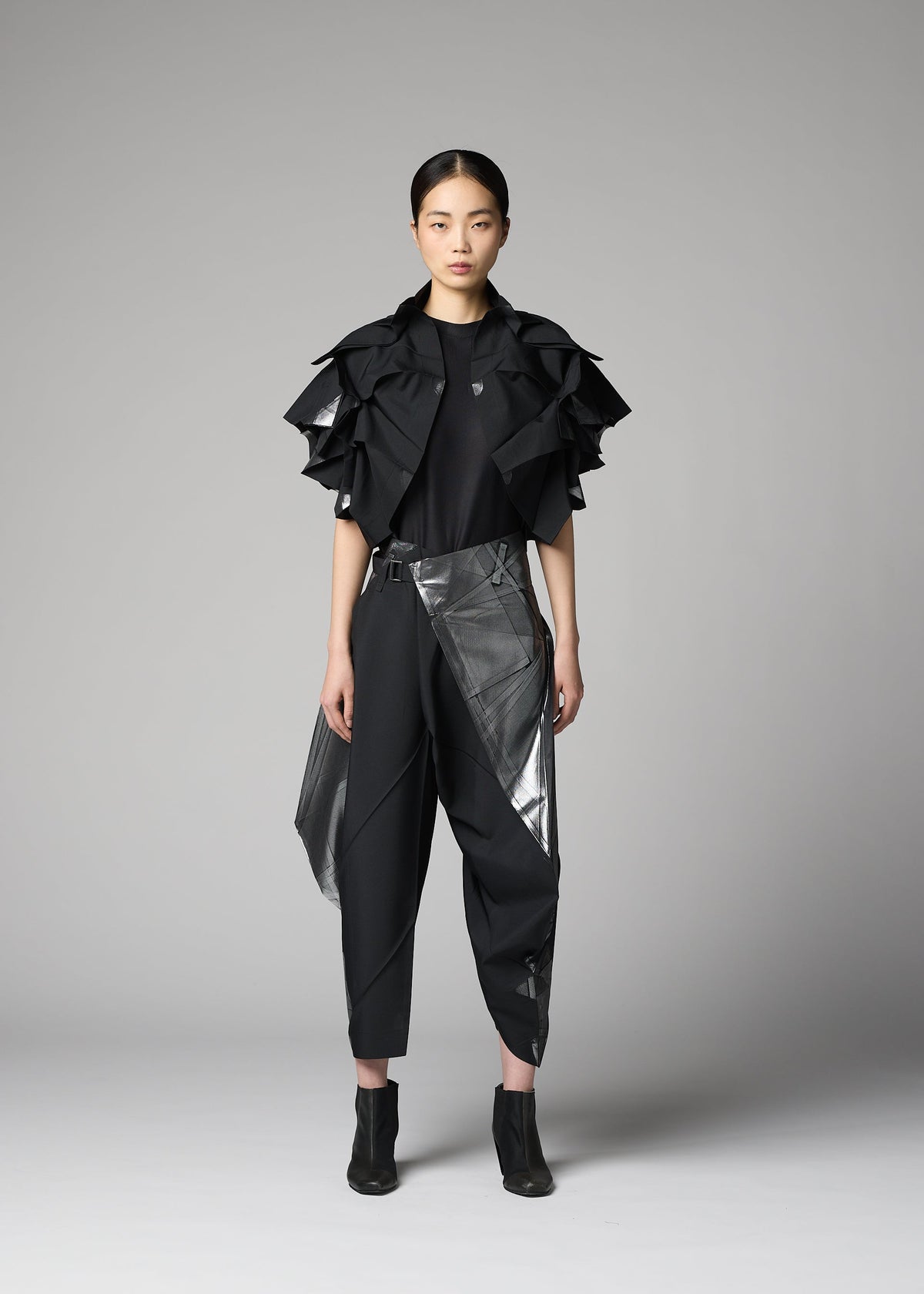 A woman faces forward against a gray background, wearing the No.10-1 jacket by 132 5. ISSEY MIYAKE: a black haori-style ruffled short-sleeve design, paired with a black t-shirt, angular-paneled pants, ankle boots, and tied-back hair. view 6 of 9