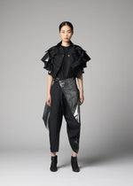 A woman faces forward against a gray background, wearing the No.10-1 jacket by 132 5. ISSEY MIYAKE: a black haori-style ruffled short-sleeve design, paired with a black t-shirt, angular-paneled pants, ankle boots, and tied-back hair.