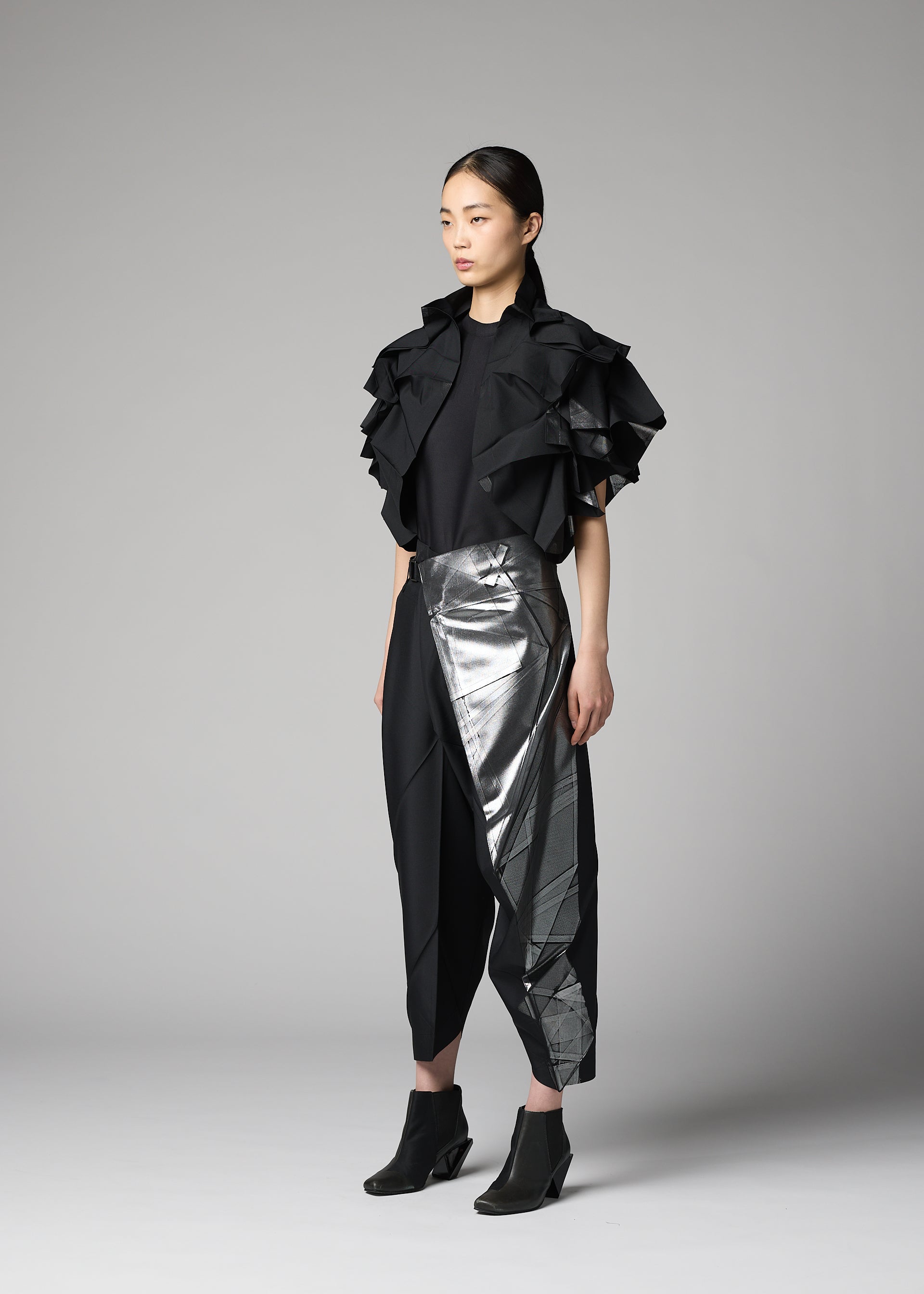 Zoomed view A woman models the No.10-1 ensemble by 132 5. ISSEY MIYAKE: a black top with bold ruffled sleeves, black cropped pants, a metallic silver asymmetrical skirt overlay, a haori-style jacket, and black ankle boots against a plain background.