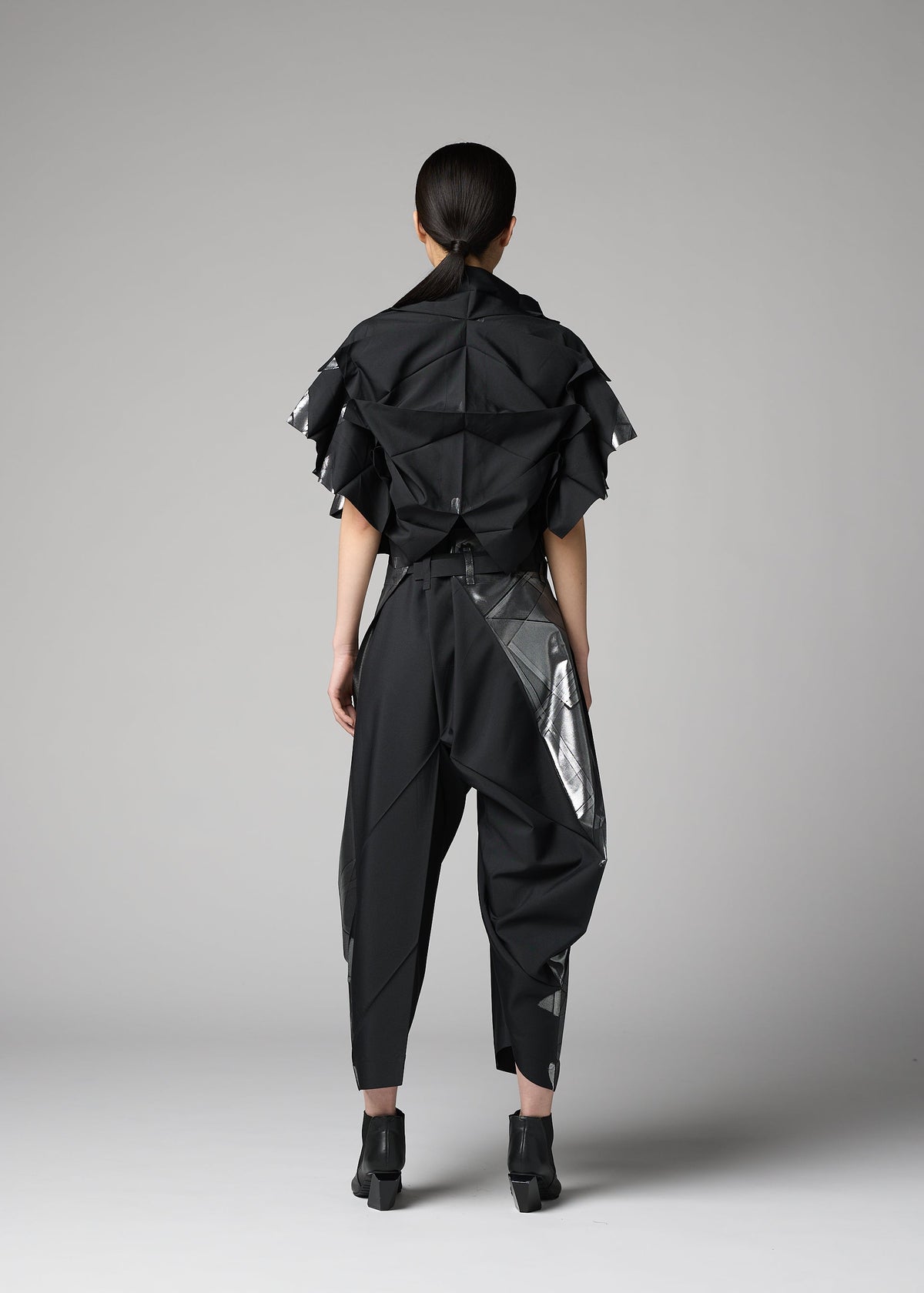 A person faces away from the camera in a plain studio, wearing 132 5. ISSEY MIYAKE's No.10-1, an avant-garde haori-style jacket with structured, layered details paired with pants featuring shiny silver accents. view 8 of 9