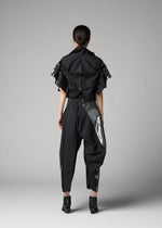 A person faces away from the camera in a plain studio, wearing 132 5. ISSEY MIYAKE's No.10-1, an avant-garde haori-style jacket with structured, layered details paired with pants featuring shiny silver accents.