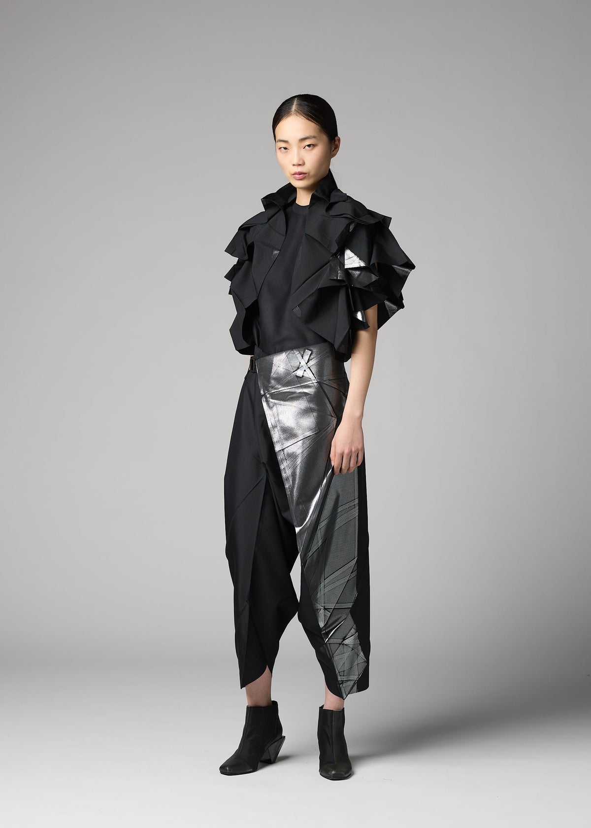 A woman poses against a gray background in the No.10-1 outfit by 132 5. ISSEY MIYAKE, featuring ruffled short sleeves, asymmetrical metallic silver accents on skirt-like pants, a foil-embellished jacket, and black ankle boots. view 9 of 9