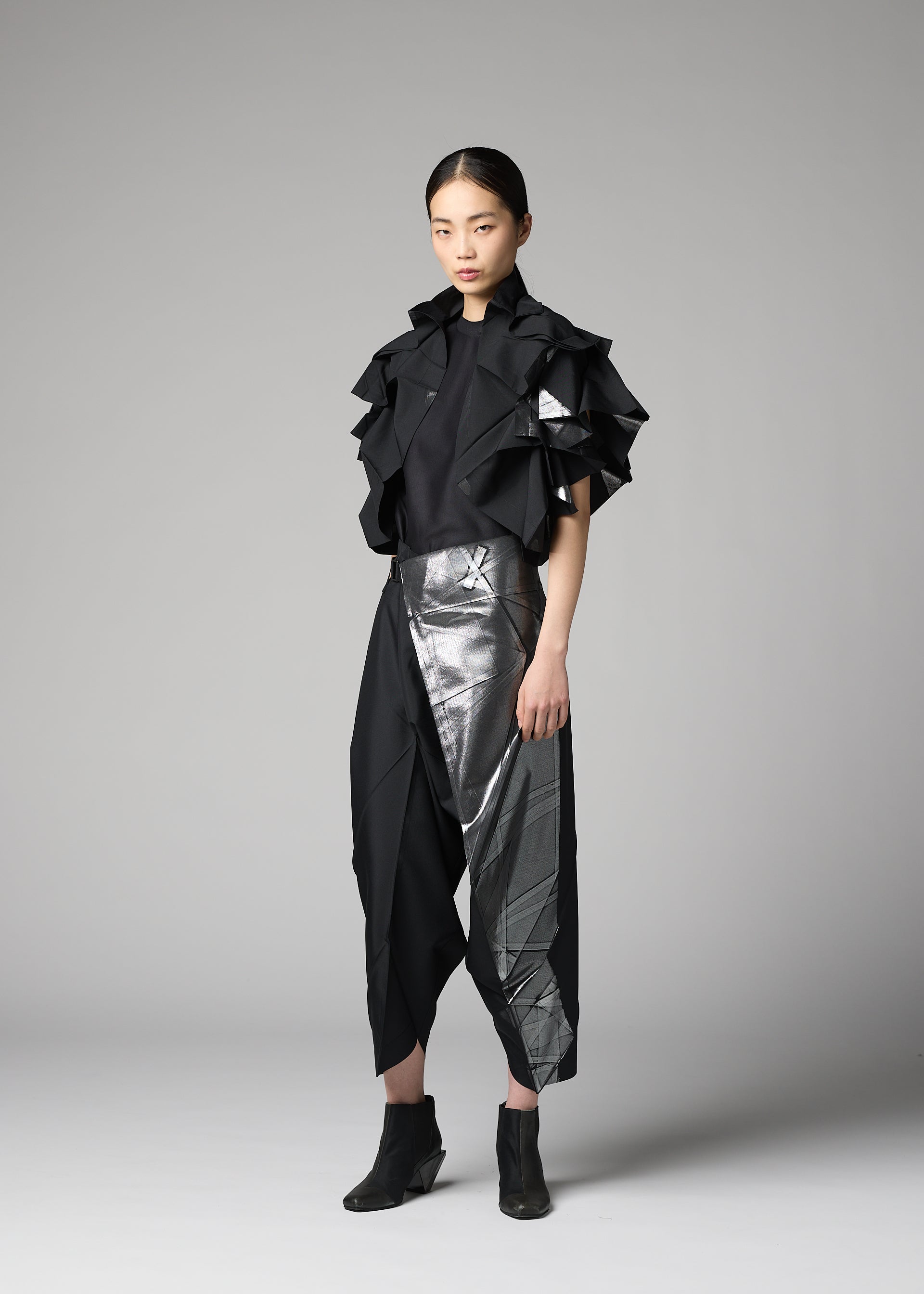 Zoomed view A woman poses against a gray background in the No.10-1 outfit by 132 5. ISSEY MIYAKE, featuring ruffled short sleeves, asymmetrical metallic silver accents on skirt-like pants, a foil-embellished jacket, and black ankle boots.