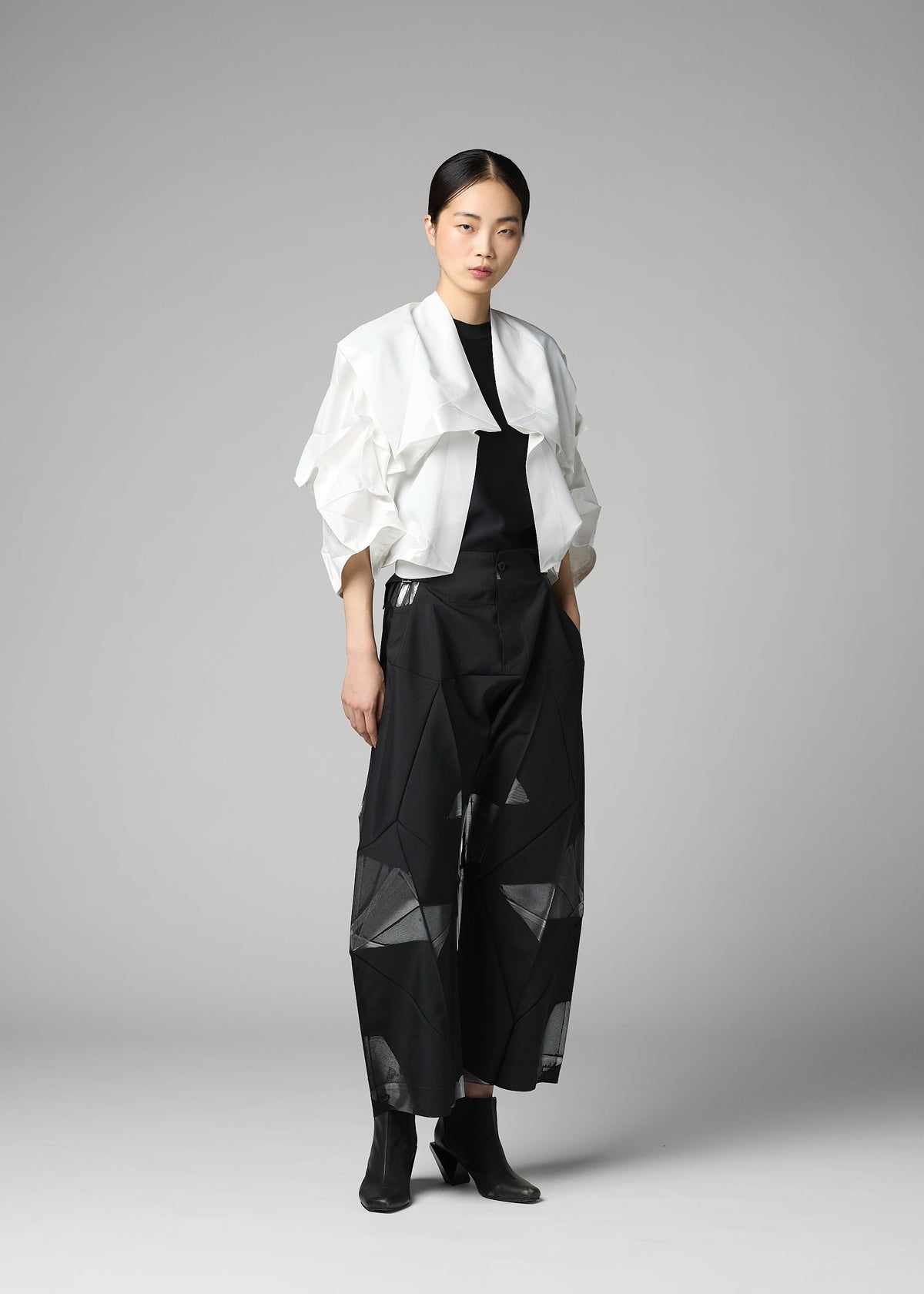A woman stands against a gray background wearing the No.1-2 wide black pants with geometric patterns, crafted from recycled polyester by 132 5. ISSEY MIYAKE, styled with a white cropped haori jacket featuring ruffled sleeves, a black top, and black ankle boots. view 6 of 9