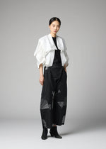A woman stands against a gray background wearing the No.1-2 wide black pants with geometric patterns, crafted from recycled polyester by 132 5. ISSEY MIYAKE, styled with a white cropped haori jacket featuring ruffled sleeves, a black top, and black ankle boots.