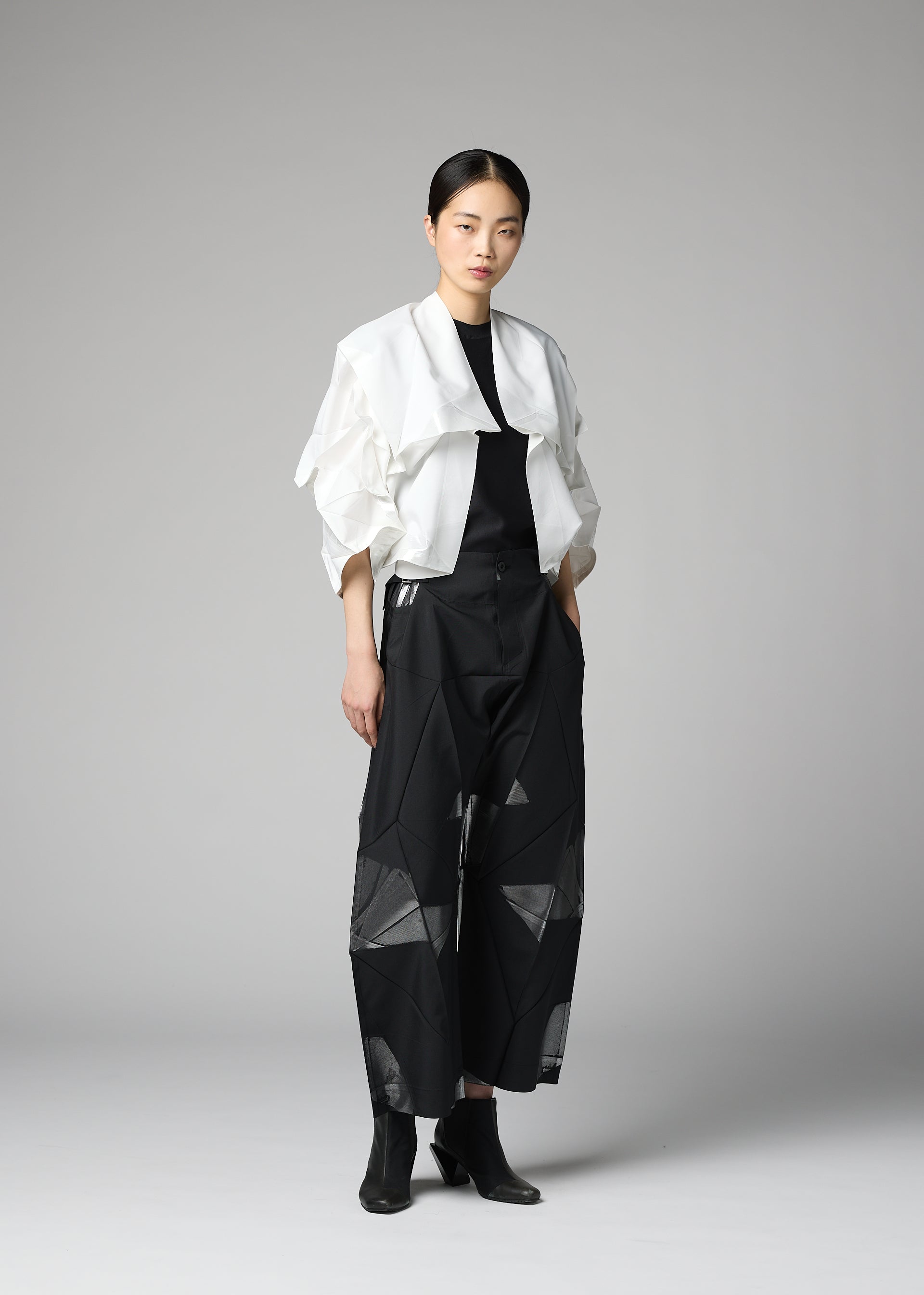 Zoomed view A woman stands against a gray background wearing the No.1-2 wide black pants with geometric patterns, crafted from recycled polyester by 132 5. ISSEY MIYAKE, styled with a white cropped haori jacket featuring ruffled sleeves, a black top, and black ankle boots.