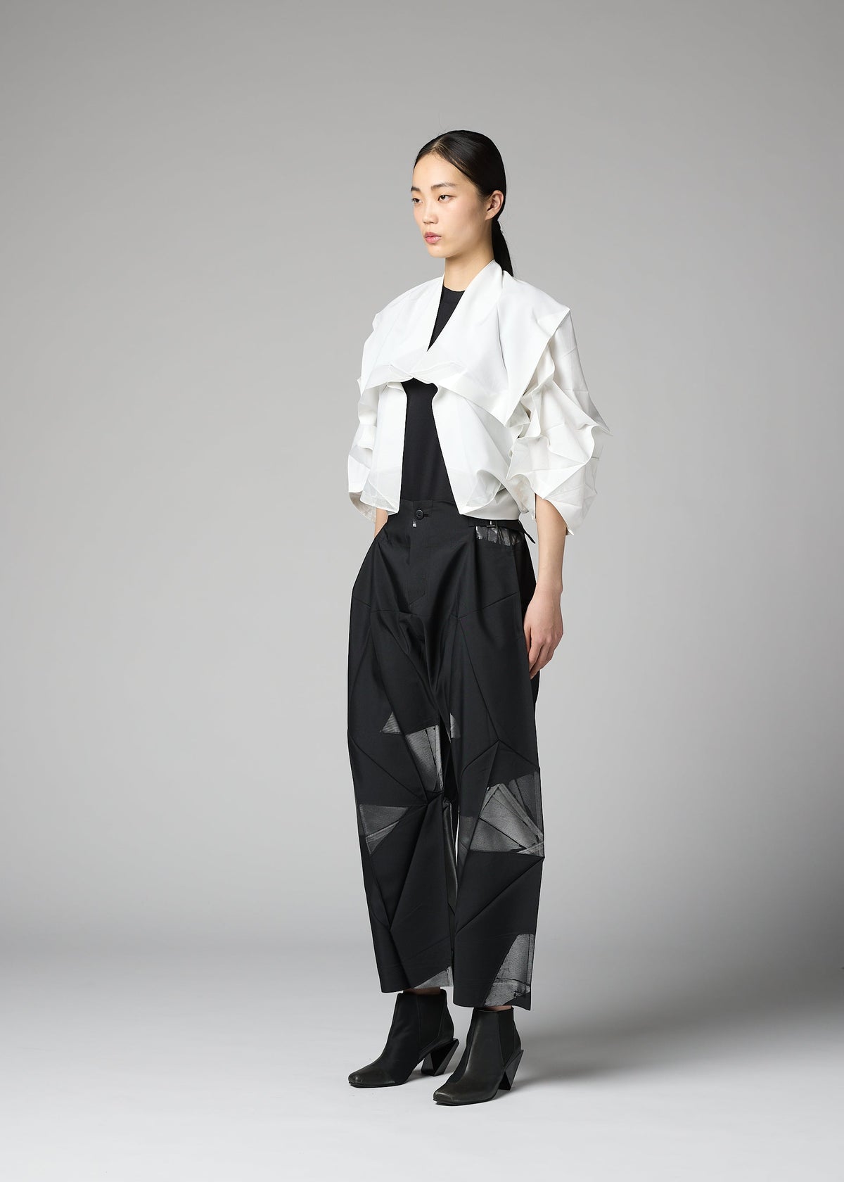 A woman stands in a studio against a plain gray background, wearing the No.1-2 structured white haori jacket by 132 5. ISSEY MIYAKE over a black top and wide-leg black pants with geometric cutouts. The jacket features subtle foil crimped details that add a modern edge to her look, which is completed with black ankle boots. view 7 of 9