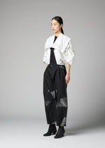 A woman stands in a studio against a plain gray background, wearing the No.1-2 structured white haori jacket by 132 5. ISSEY MIYAKE over a black top and wide-leg black pants with geometric cutouts. The jacket features subtle foil crimped details that add a modern edge to her look, which is completed with black ankle boots.