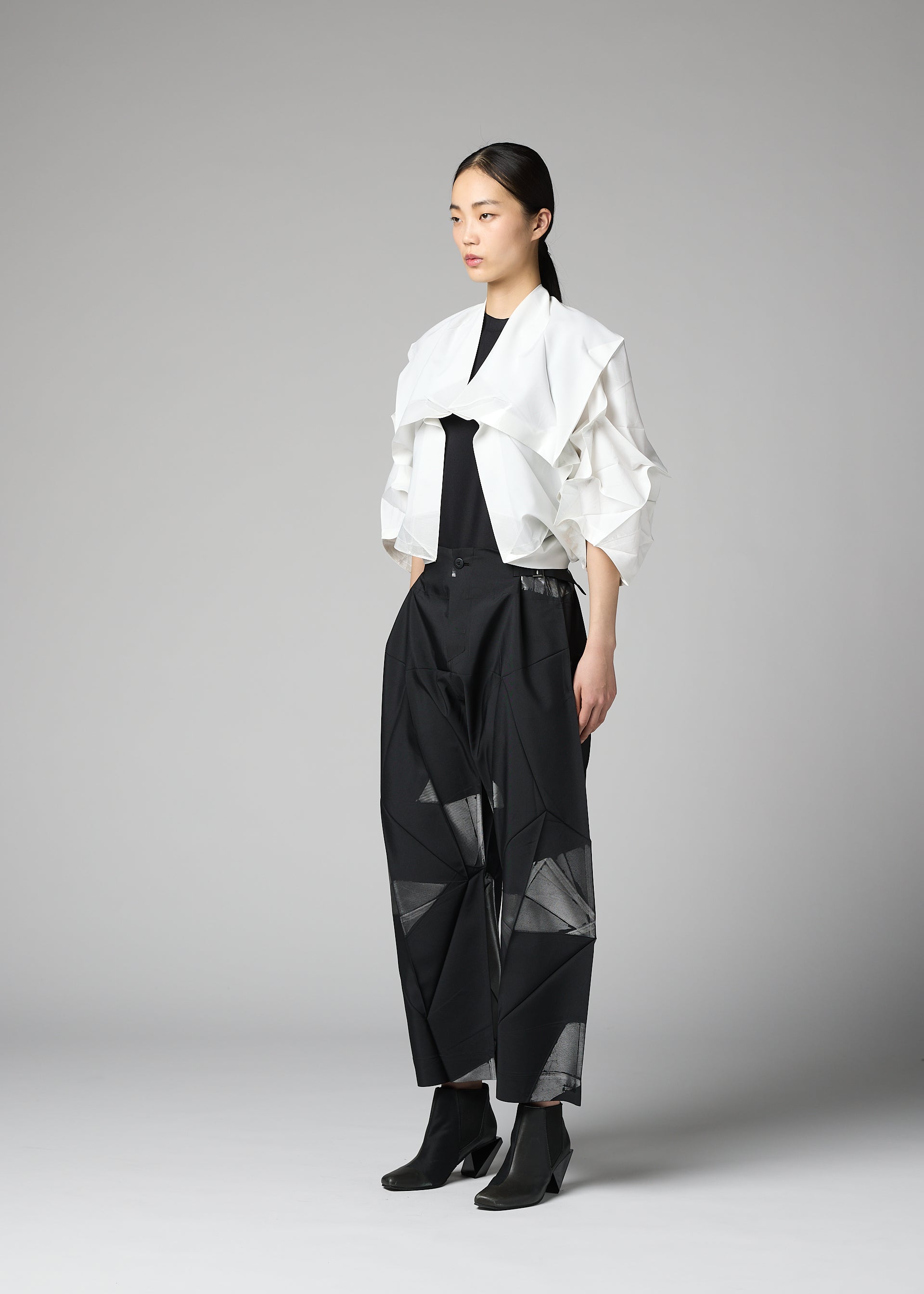 Zoomed view A woman stands in a studio against a plain gray background, wearing the No.1-2 structured white haori jacket by 132 5. ISSEY MIYAKE over a black top and wide-leg black pants with geometric cutouts. The jacket features subtle foil crimped details that add a modern edge to her look, which is completed with black ankle boots.