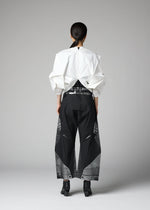 A person stands facing away against a plain gray background, dressed in the 132 5. ISSEY MIYAKE No.1-2 top—a sculptural white garment featuring geometric folds crafted from recycled polyester—paired with loose black pants with sheer panels, a shiny belt, and black ankle boots.
