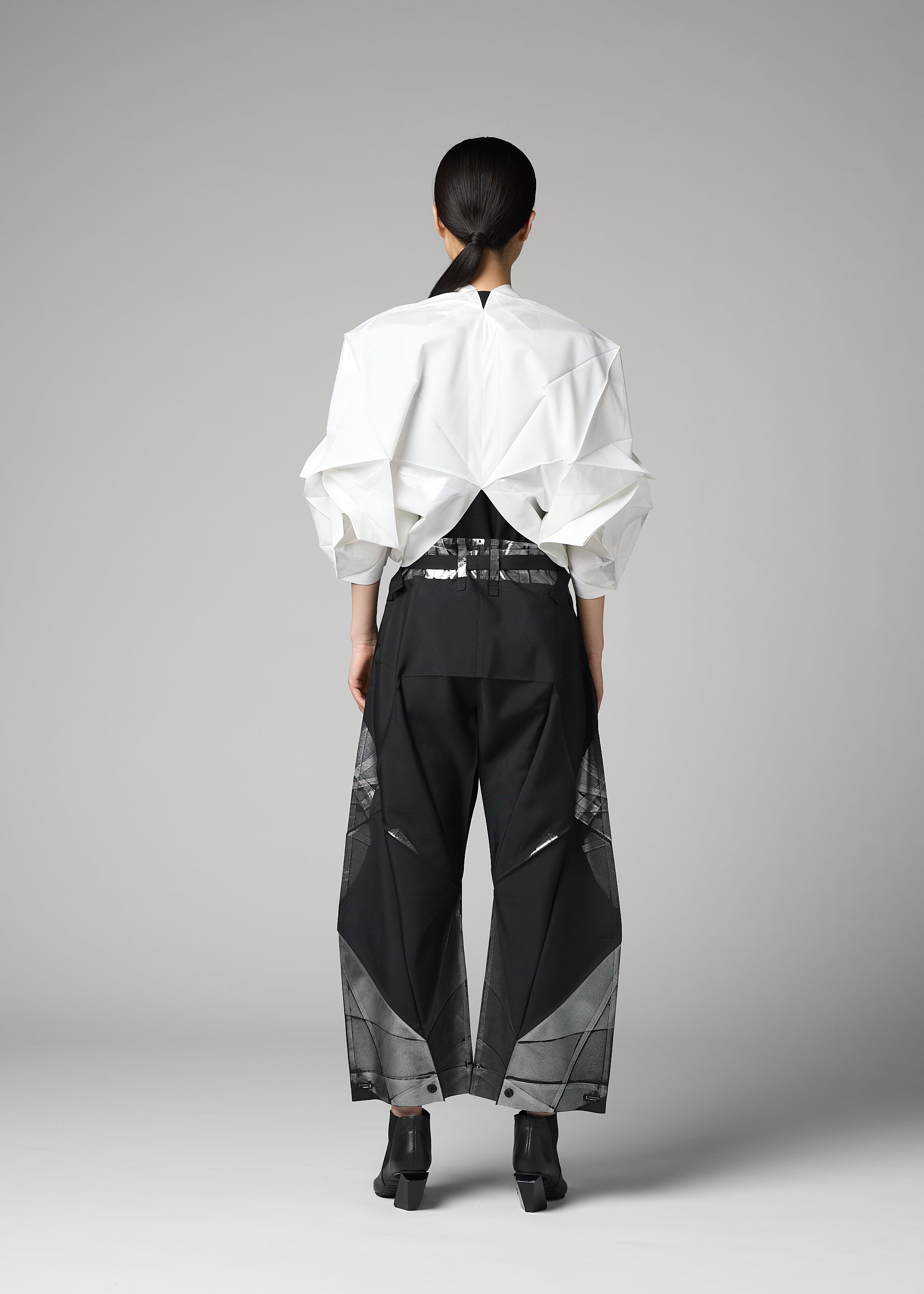 Zoomed view A person stands facing away against a plain gray background, dressed in the 132 5. ISSEY MIYAKE No.1-2 top—a sculptural white garment featuring geometric folds crafted from recycled polyester—paired with loose black pants with sheer panels, a shiny belt, and black ankle boots.
