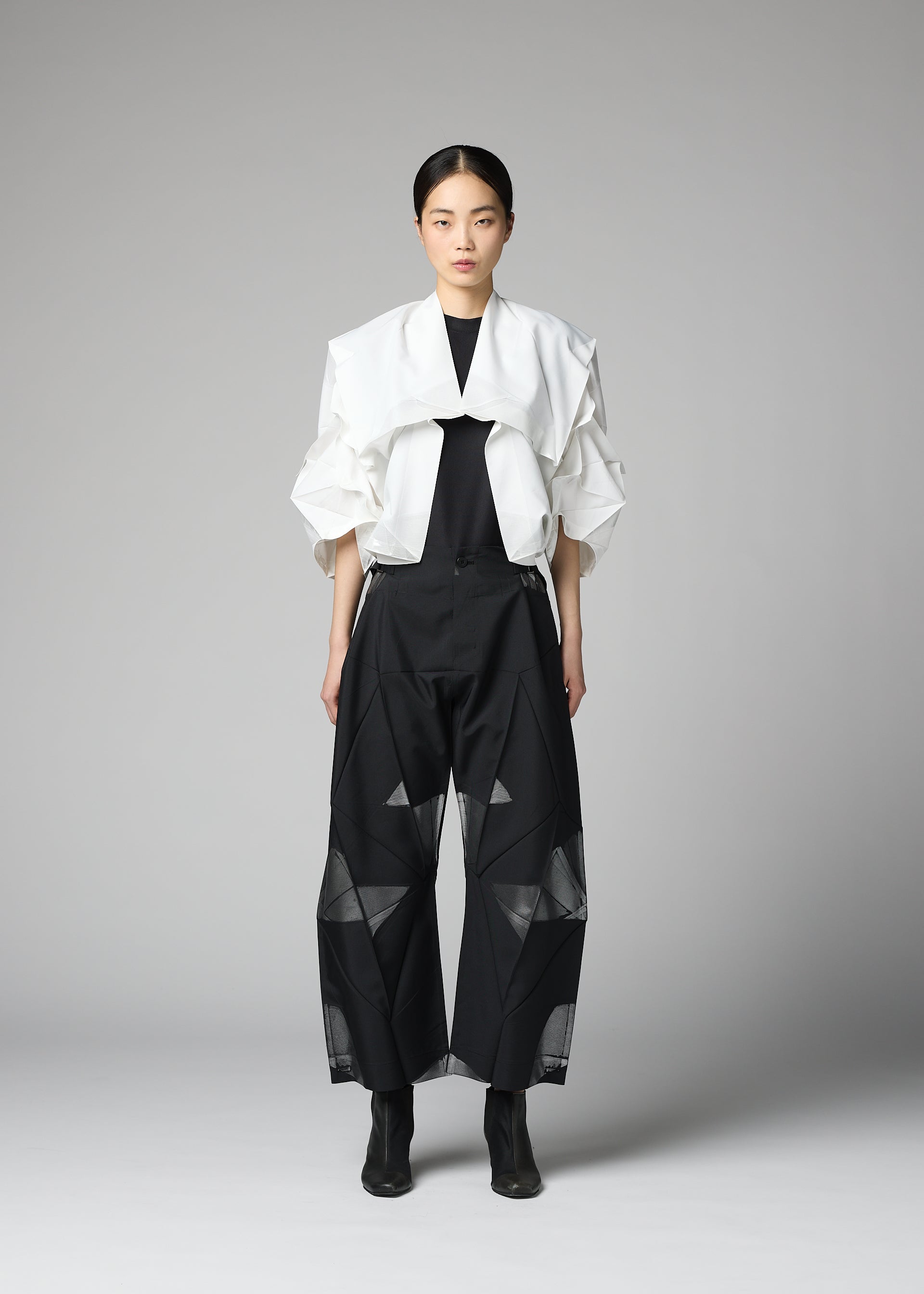 Zoomed view A person stands against a plain gray background wearing the No.1-2 haori jacket by 132 5. ISSEY MIYAKE—crafted from recycled polyester in white—layered over a black top and wide black pants with sheer geometric panels, paired with black ankle boots.