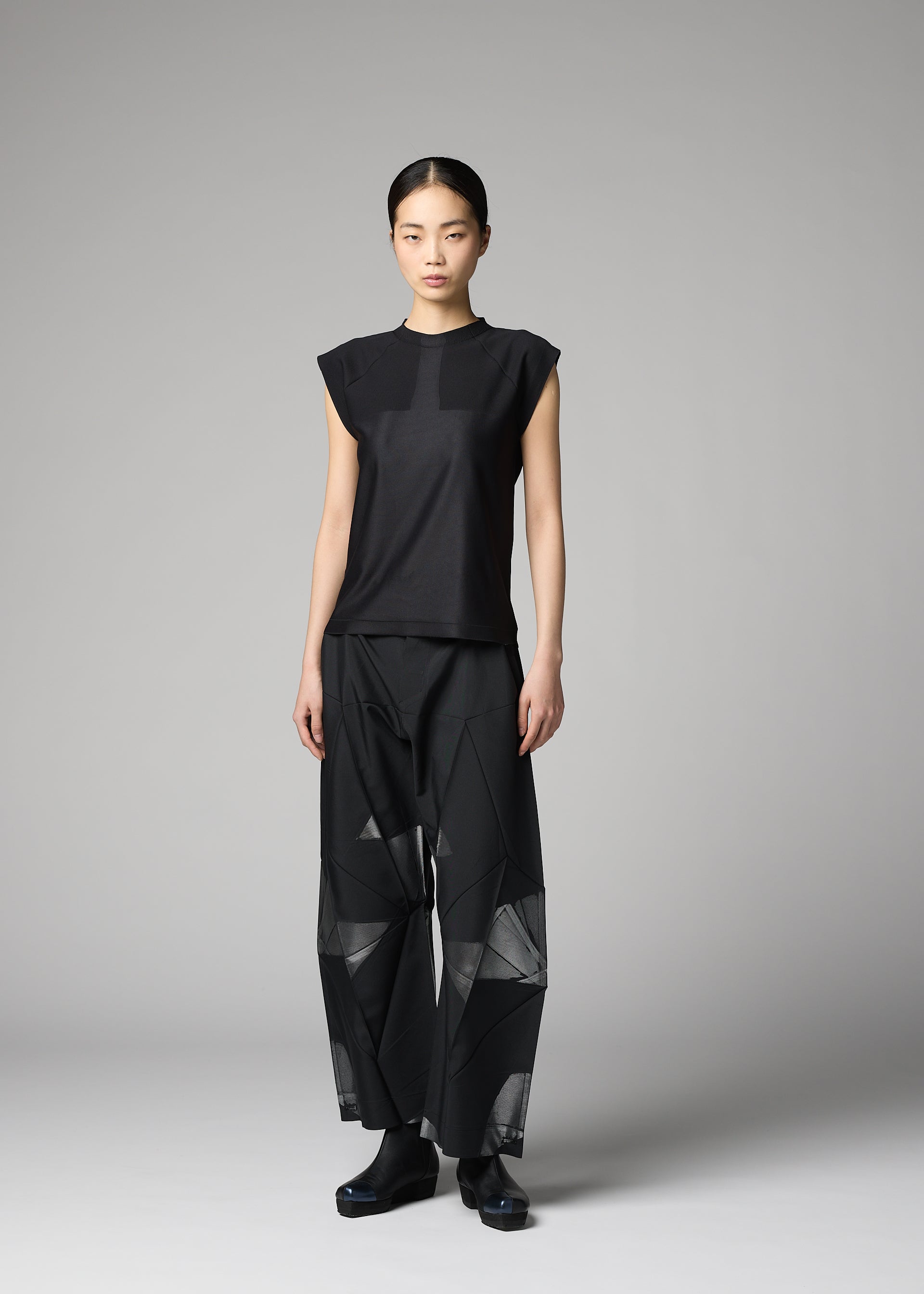 Zoomed view A person with straight dark hair tied back models the F-KN black sleeveless top and wide pants with luster, folds, and geometric sheer panels by 132 5. ISSEY MIYAKE, standing against a plain gray background.