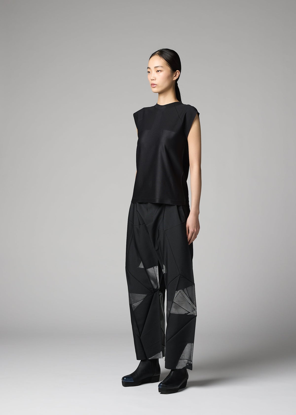 A woman models the F-KN ensemble by 132 5. ISSEY MIYAKE—a sleeveless black top and loose, geometric-patterned pants with a subtle sheen—standing in a studio against a gray background, her hair pulled back as she gazes to the side. view 5 of 6