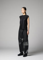 A woman models the F-KN ensemble by 132 5. ISSEY MIYAKE—a sleeveless black top and loose, geometric-patterned pants with a subtle sheen—standing in a studio against a gray background, her hair pulled back as she gazes to the side.