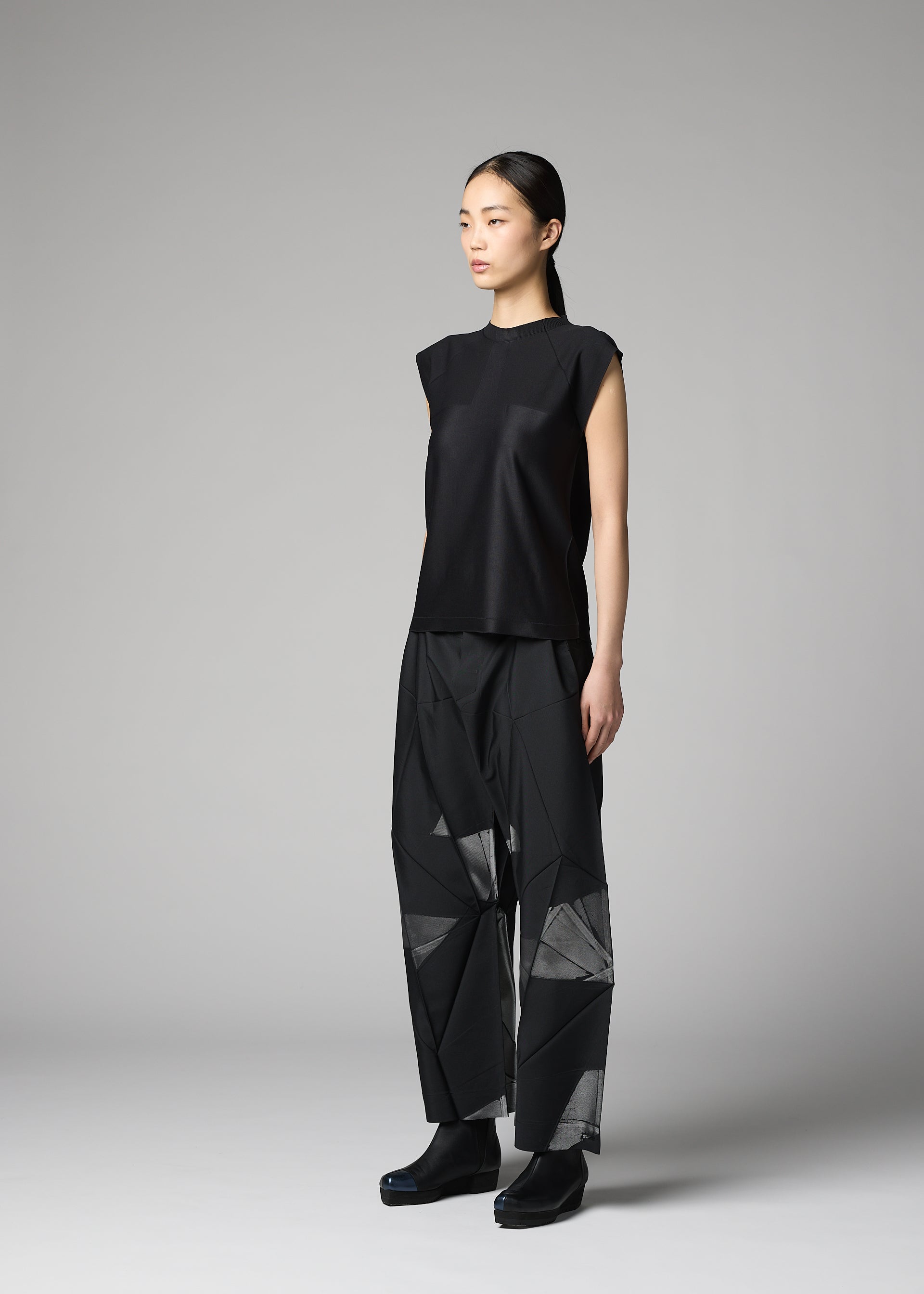 Zoomed view A woman models the F-KN ensemble by 132 5. ISSEY MIYAKE—a sleeveless black top and loose, geometric-patterned pants with a subtle sheen—standing in a studio against a gray background, her hair pulled back as she gazes to the side.
