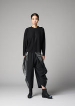 A woman stands in a studio against a plain gray background, wearing the F-KN set by 132 5. ISSEY MIYAKE—loose black long-sleeve top and wide-leg pants with shiny, textured panels—paired with black ankle boots.