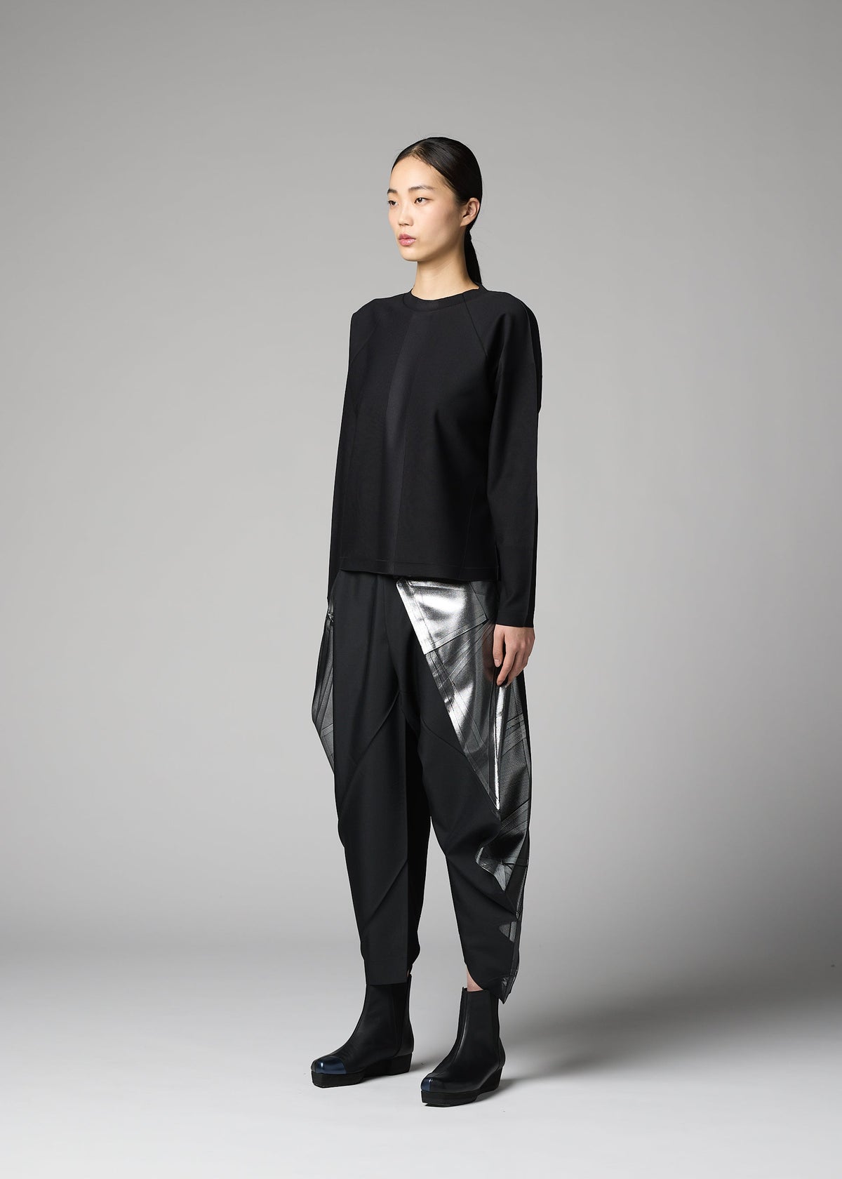 A woman stands in a studio against a plain gray background, wearing the F-KN top and geometric black and silver pants by 132 5. ISSEY MIYAKE, their folds hinting at technique 132.5, paired with black ankle boots. view 5 of 6