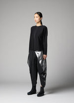A woman stands in a studio against a plain gray background, wearing the F-KN top and geometric black and silver pants by 132 5. ISSEY MIYAKE, their folds hinting at technique 132.5, paired with black ankle boots.