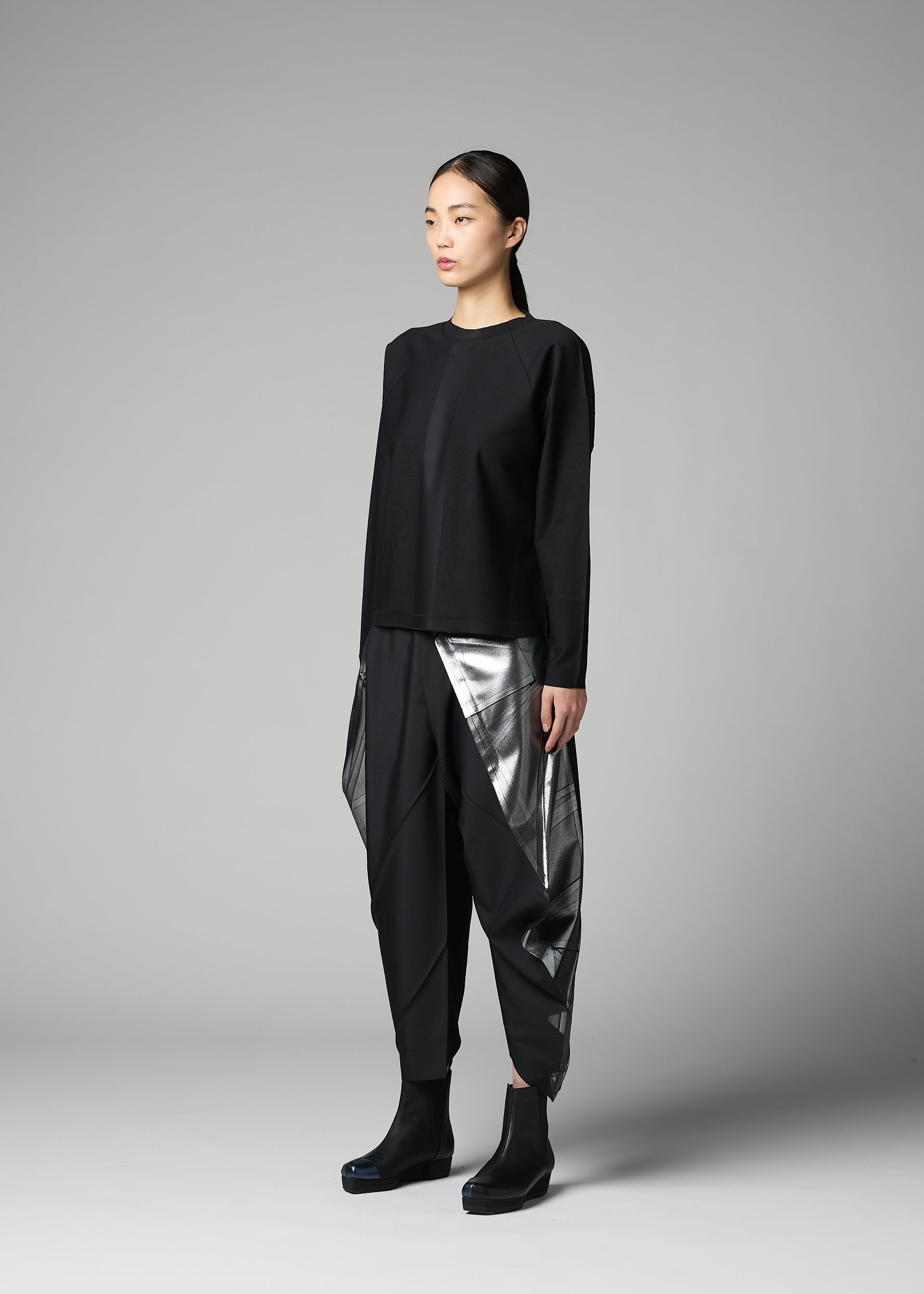 Zoomed view A woman stands in a studio against a plain gray background, wearing the F-KN top and geometric black and silver pants by 132 5. ISSEY MIYAKE, their folds hinting at technique 132.5, paired with black ankle boots.