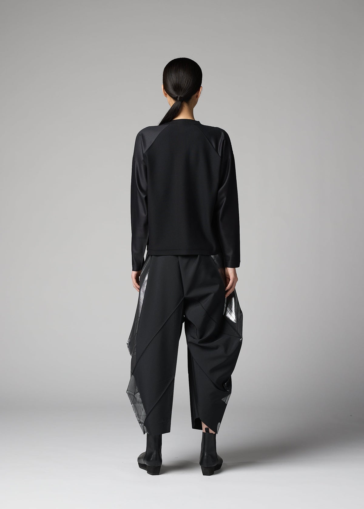 A person with long dark hair tied back stands facing away in a plain studio, wearing the F-KN black top and wide structured pants with shiny angular panels by 132 5. ISSEY MIYAKE, paired with black ankle boots. view 6 of 6