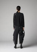A person with long dark hair tied back stands facing away in a plain studio, wearing the F-KN black top and wide structured pants with shiny angular panels by 132 5. ISSEY MIYAKE, paired with black ankle boots.